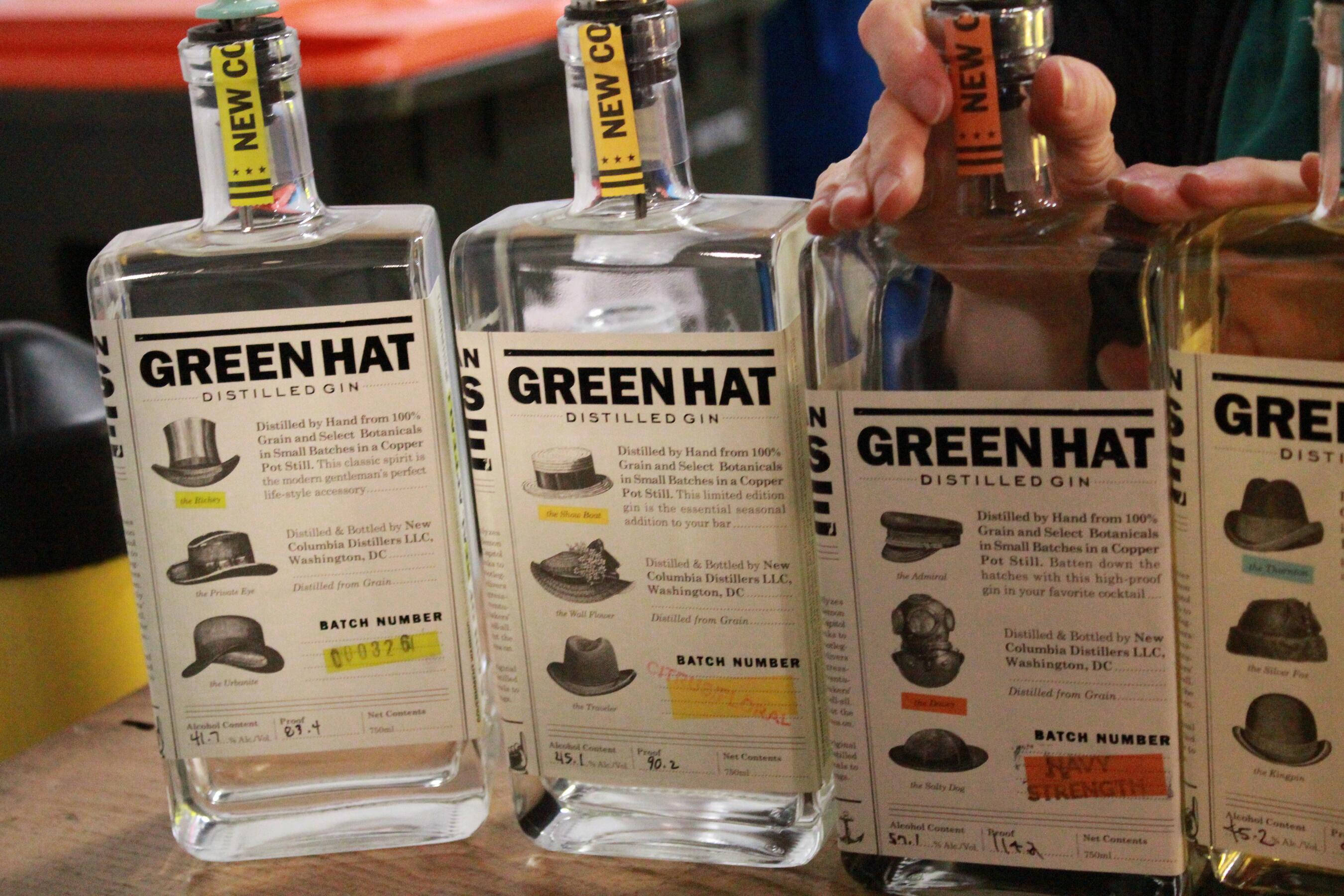 Gin 桌布 壁紙 — 四瓶透明的 Green Hat Distilled Gin 帶有獨特帽子主題標籤，放在木質表面上，其中一瓶由一隻手拿著。