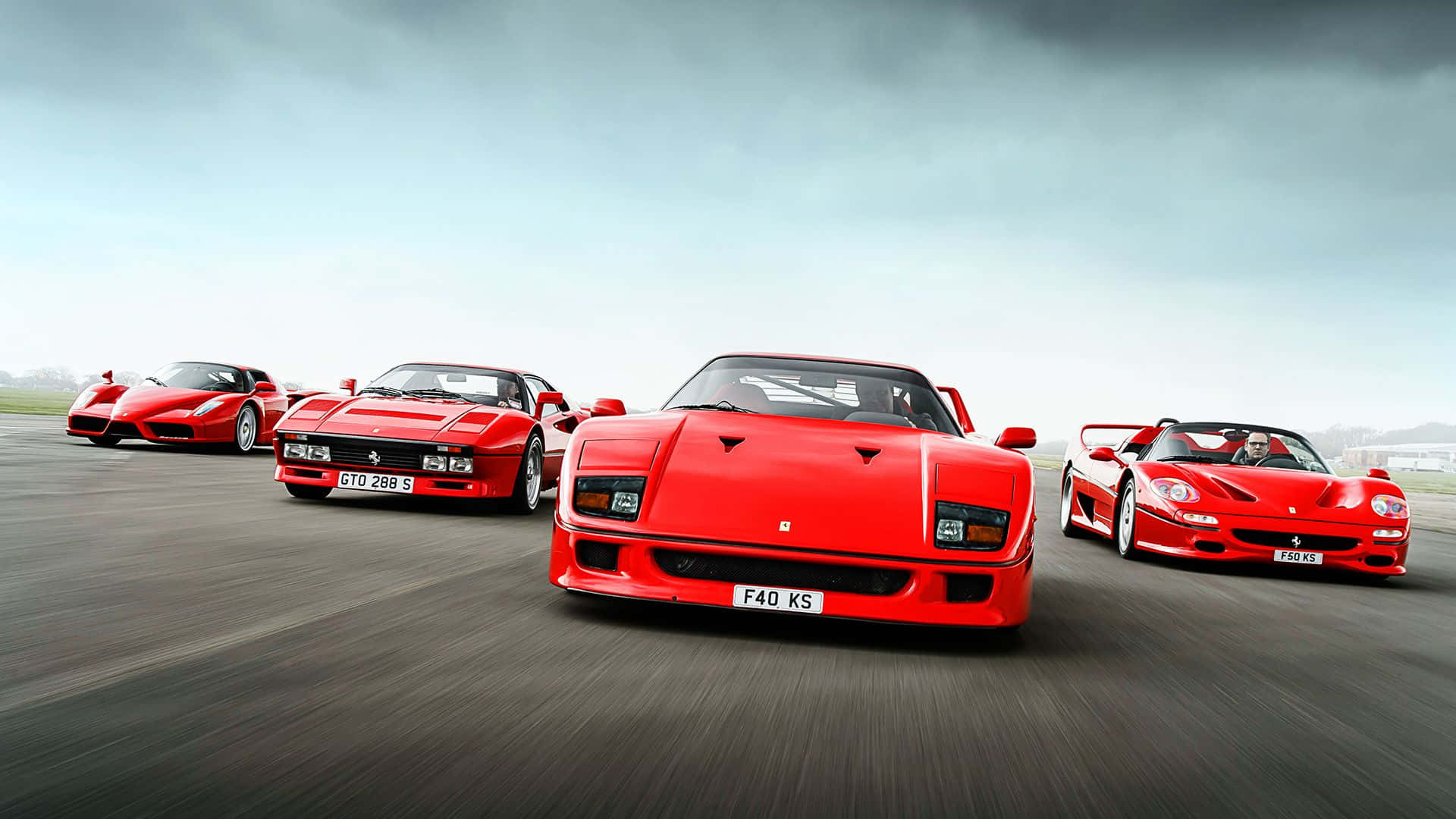 Ferrari Enzo 桌布 壁紙 — 四輛標誌性的紅色法拉利超級跑車在陰天下一條模糊的瀝青賽道上以隊形行駛。
