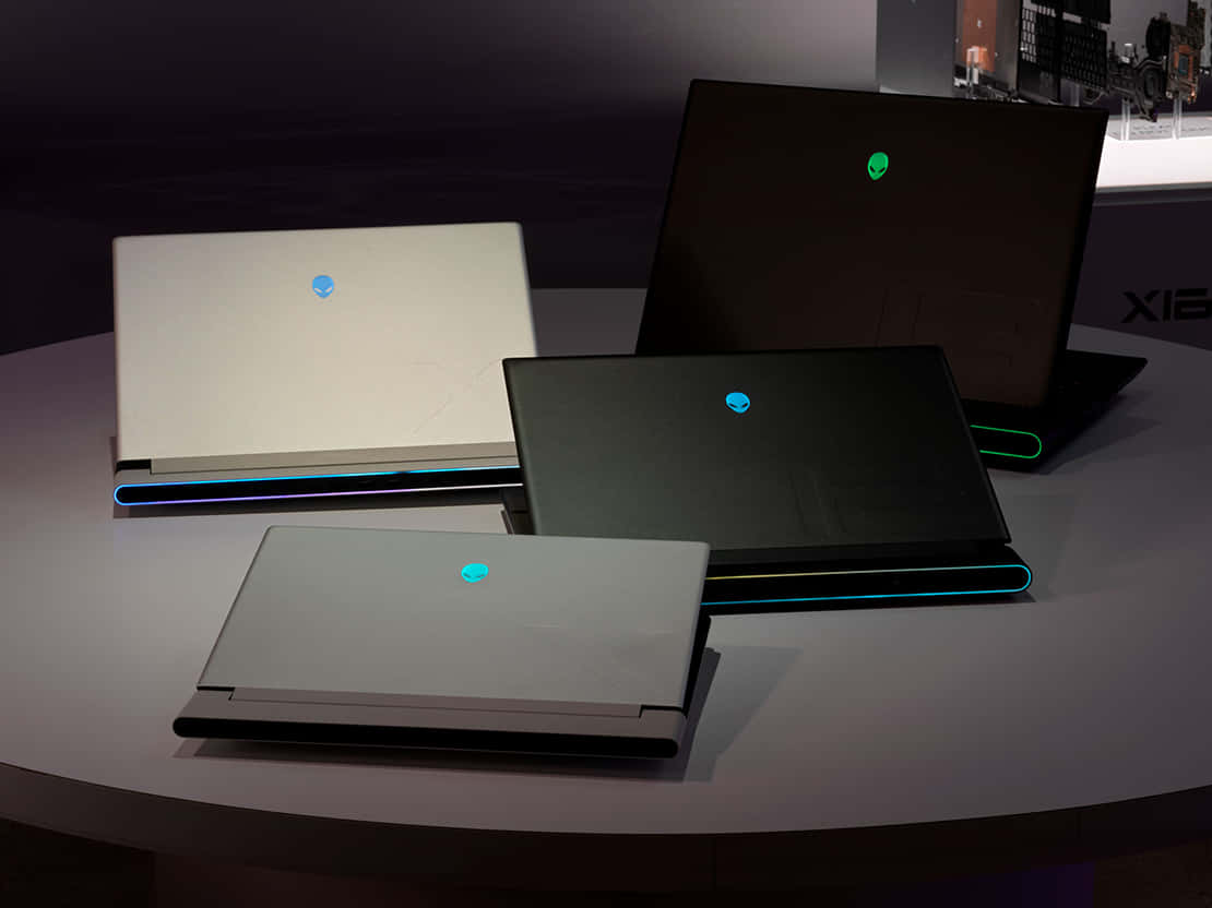 外星人 桌布 壁紙 — 四部 Alienware 遊戲筆記型電腦，帶有發光外星人標誌，展示在弧形桌上，光線昏暗。