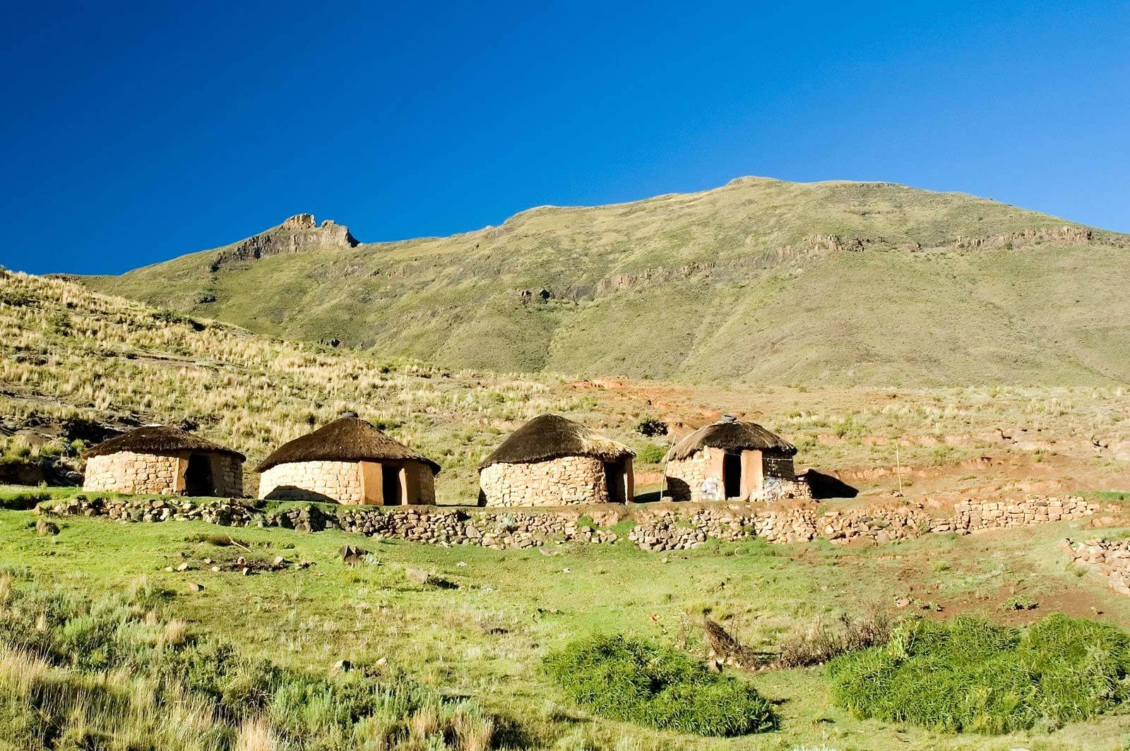 Lesotho 桌布 壁紙 — 四間茅草屋頂的石屋坐落在陽光明媚的綠色山谷中，上方是廣闊的藍天和連綿起伏的山丘。
