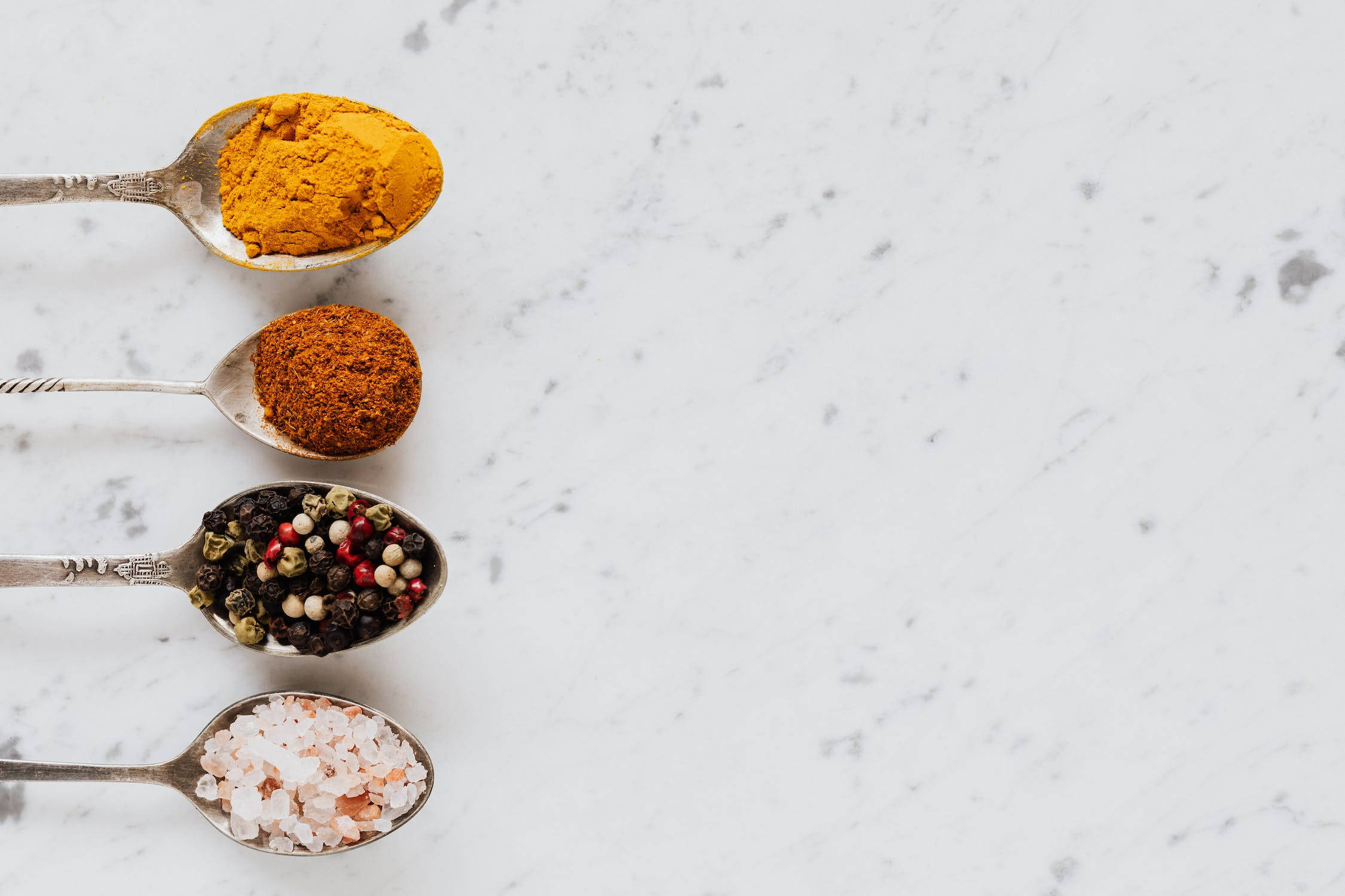 Spices 桌布 壁紙 — 四隻復古湯匙盛載著鮮豔的香料——薑黃、卡宴胡椒、黑胡椒粒和粉紅鹽——整齊地排列在白色大理石表面上。