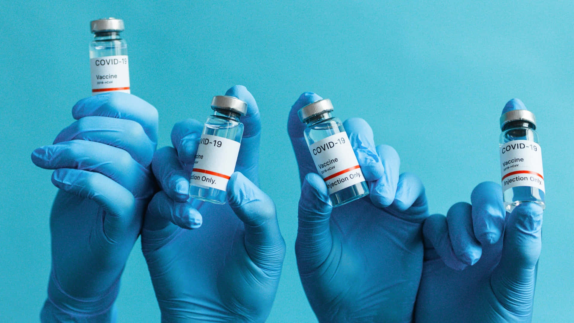 Coronavirus 桌布 壁紙 — 四隻戴手套的手在純藍色背景上持有標有COVID-19疫苗的透明小瓶。