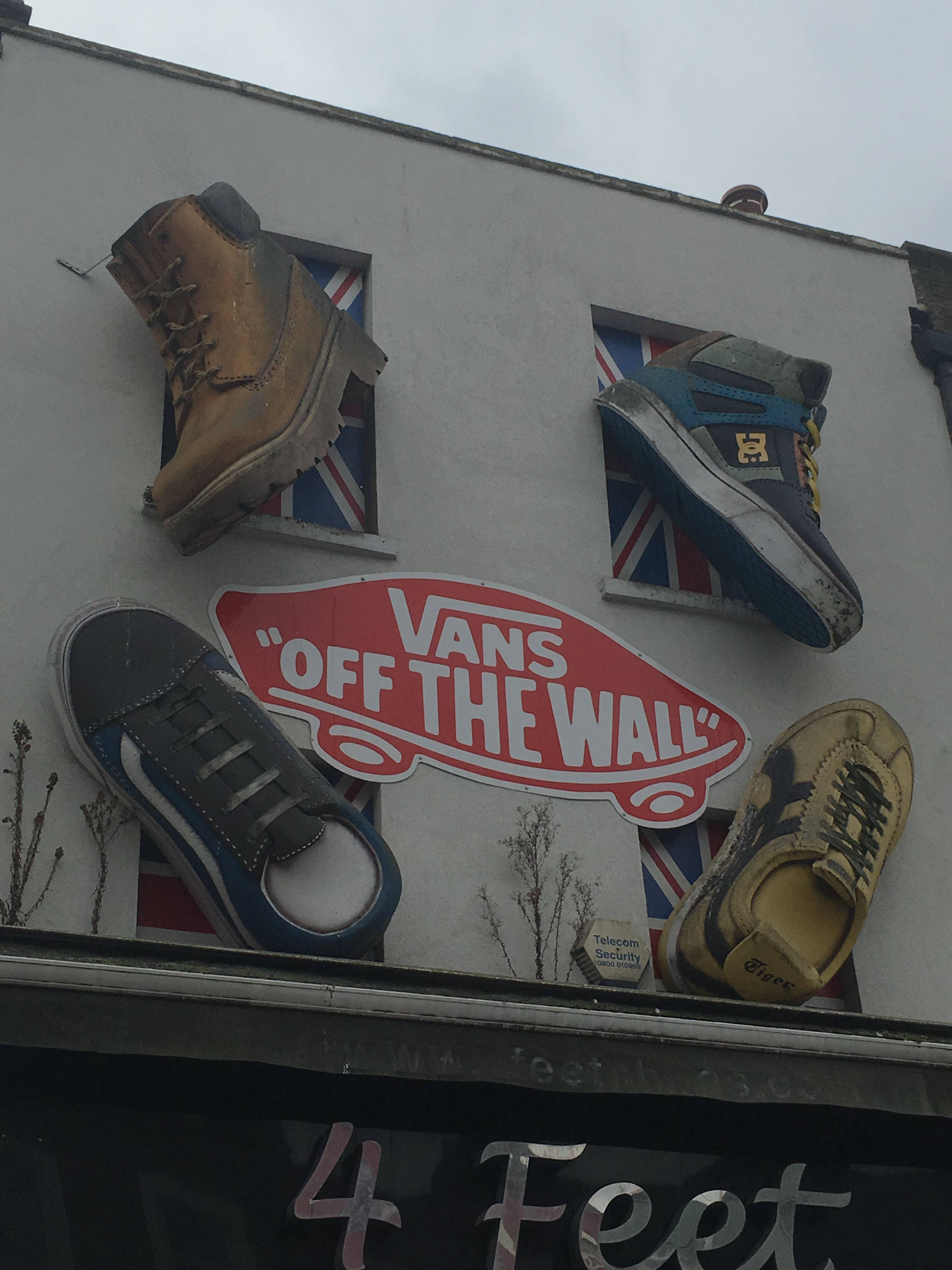 Vans Off The Wall 桌布 壁紙 — 四雙復古運動鞋和一個Vans標誌安裝在建築物外牆上，窗框為英國國旗圖案，天空多雲。