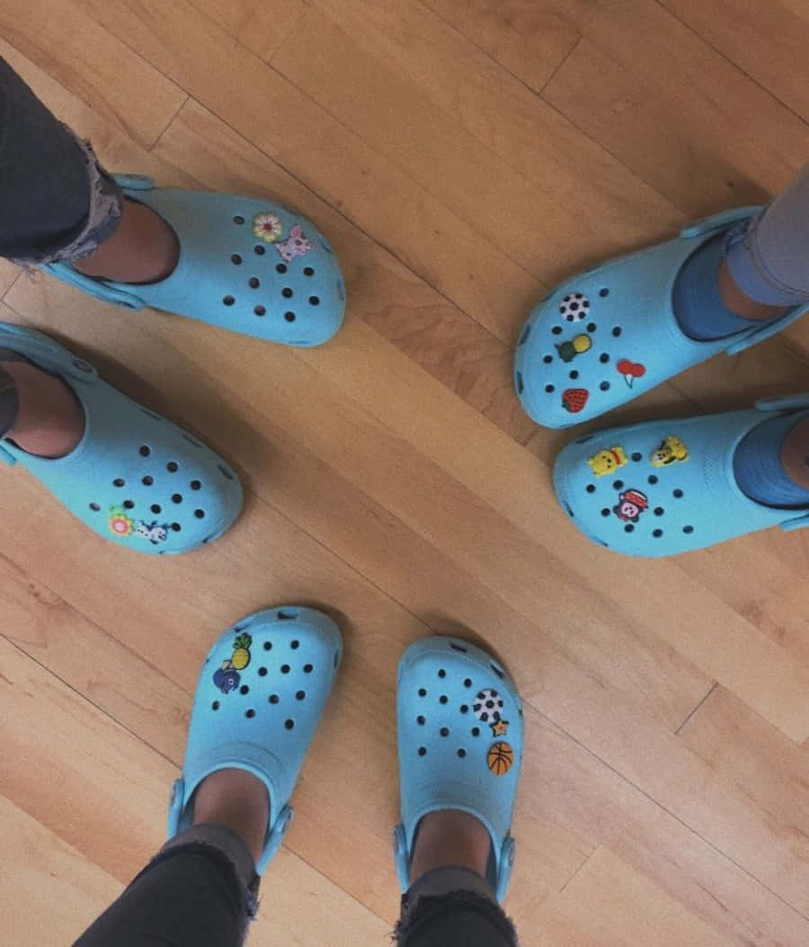 Crocs 桌布 壁紙 — 四雙淺藍色Crocs鞋上貼有色彩繽紛的卡通貼紙，從俯視角度看到它們站在溫暖的木地板上。