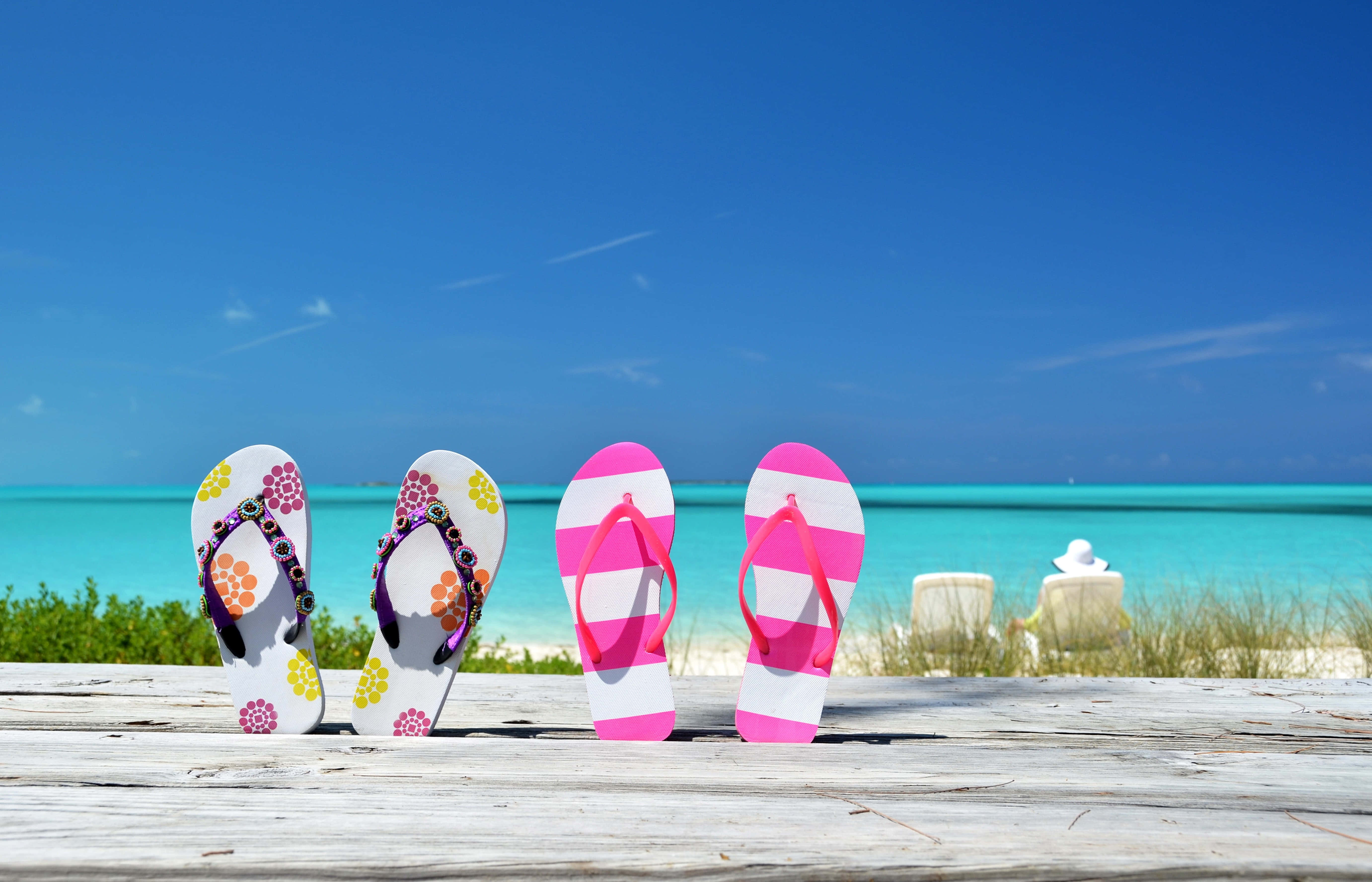 Beach Sandals 桌布 壁紙 — 四雙色彩繽紛的夾腳拖放在一個歷經風霜的木甲板上，俯瞰著清澈藍天下的碧綠海洋。