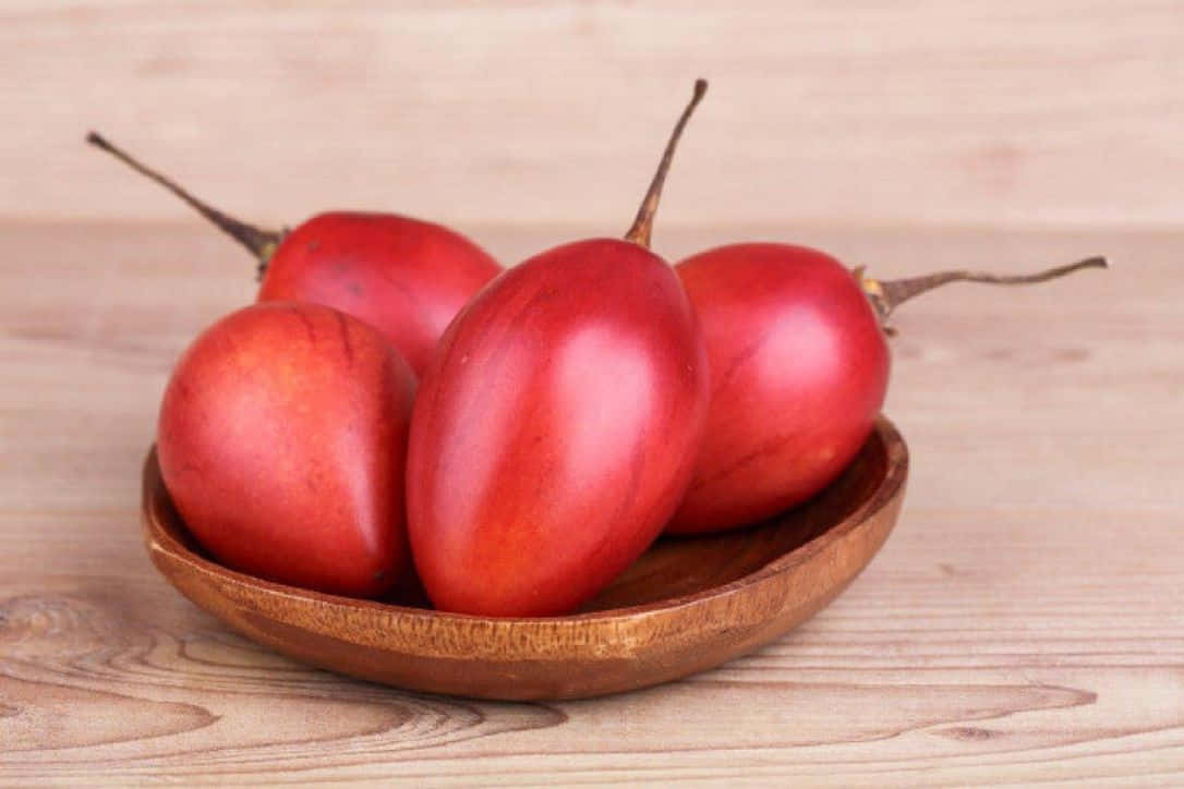 Red Fruit 桌布 壁紙 — 四顆帶莖的光澤紅茄子放在淺木碗中，置於淺色木質表面上。