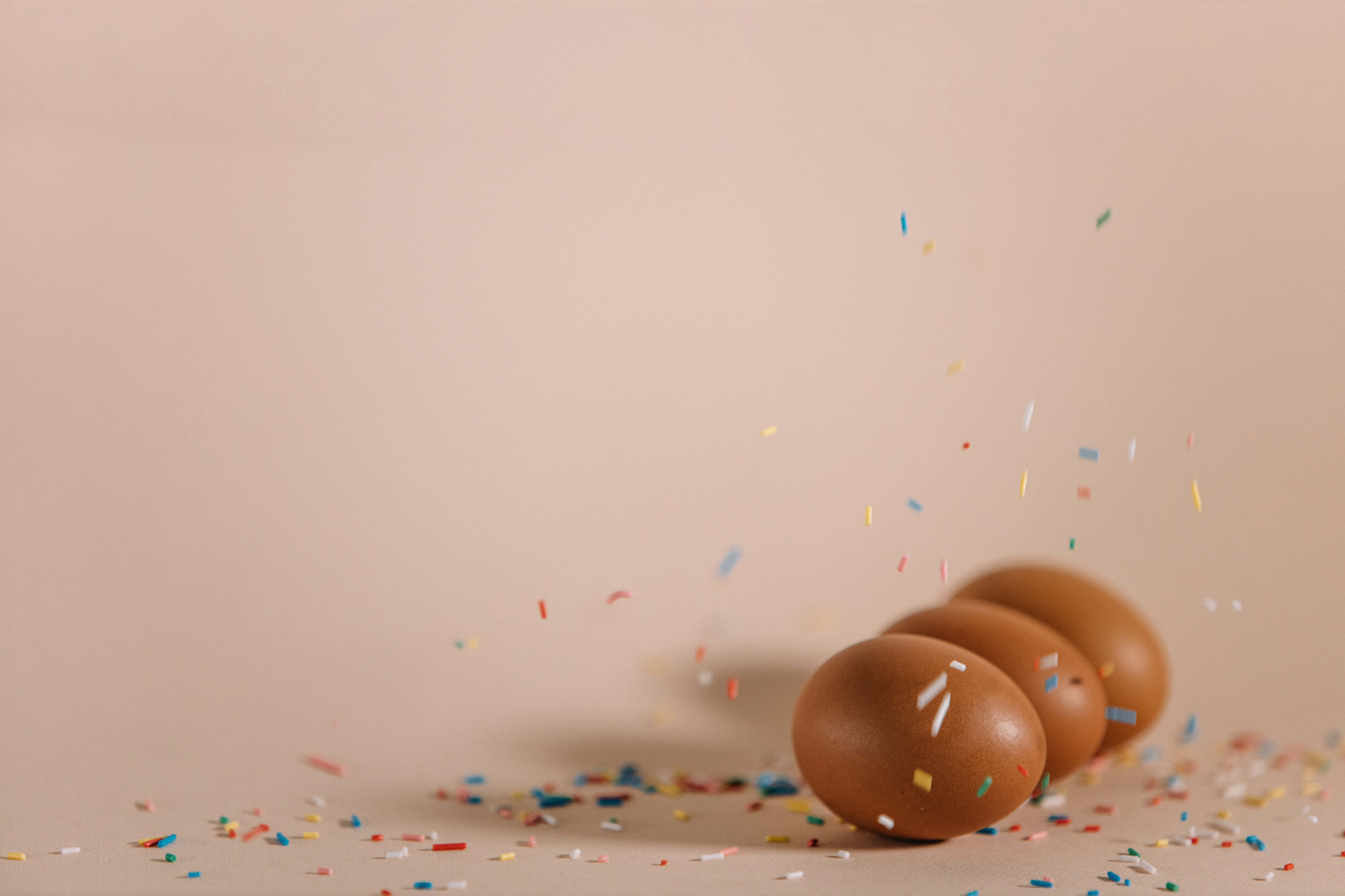 Brown Eggs 桌布 壁紙 — 四顆棕色雞蛋在米色表面上滾動，彩色紙屑以慢動作在它們周圍爆開。