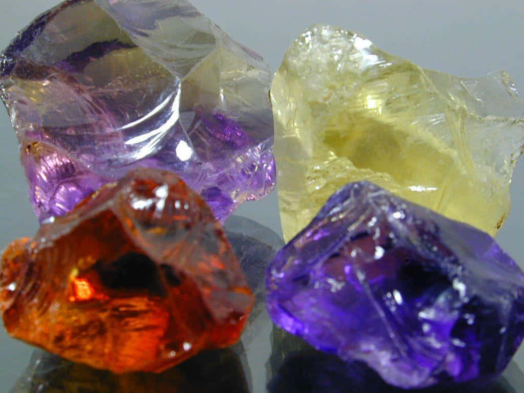 Gemstone 桌布 壁紙 — 四顆粗糙切割的寶石，呈現鮮豔的紫色、橙色和黃色色調，在反光表面上閃耀著內部反射。