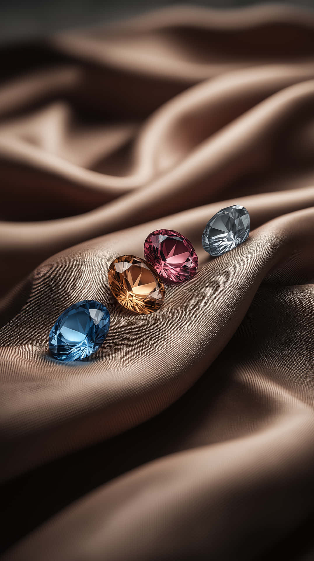 Silken Gem Assortment 壁紙 — 四顆藍色、琥珀色、粉紅色和灰色的刻面寶石，放置在帶有柔和光線的棕色緞面布料上。