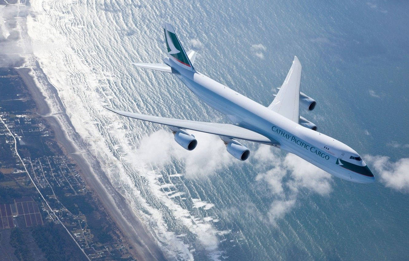 飛機 桌布 壁紙 — 國泰航空貨運珍寶機在海岸景觀上空翱翔，海浪拍岸，雲層在下方。