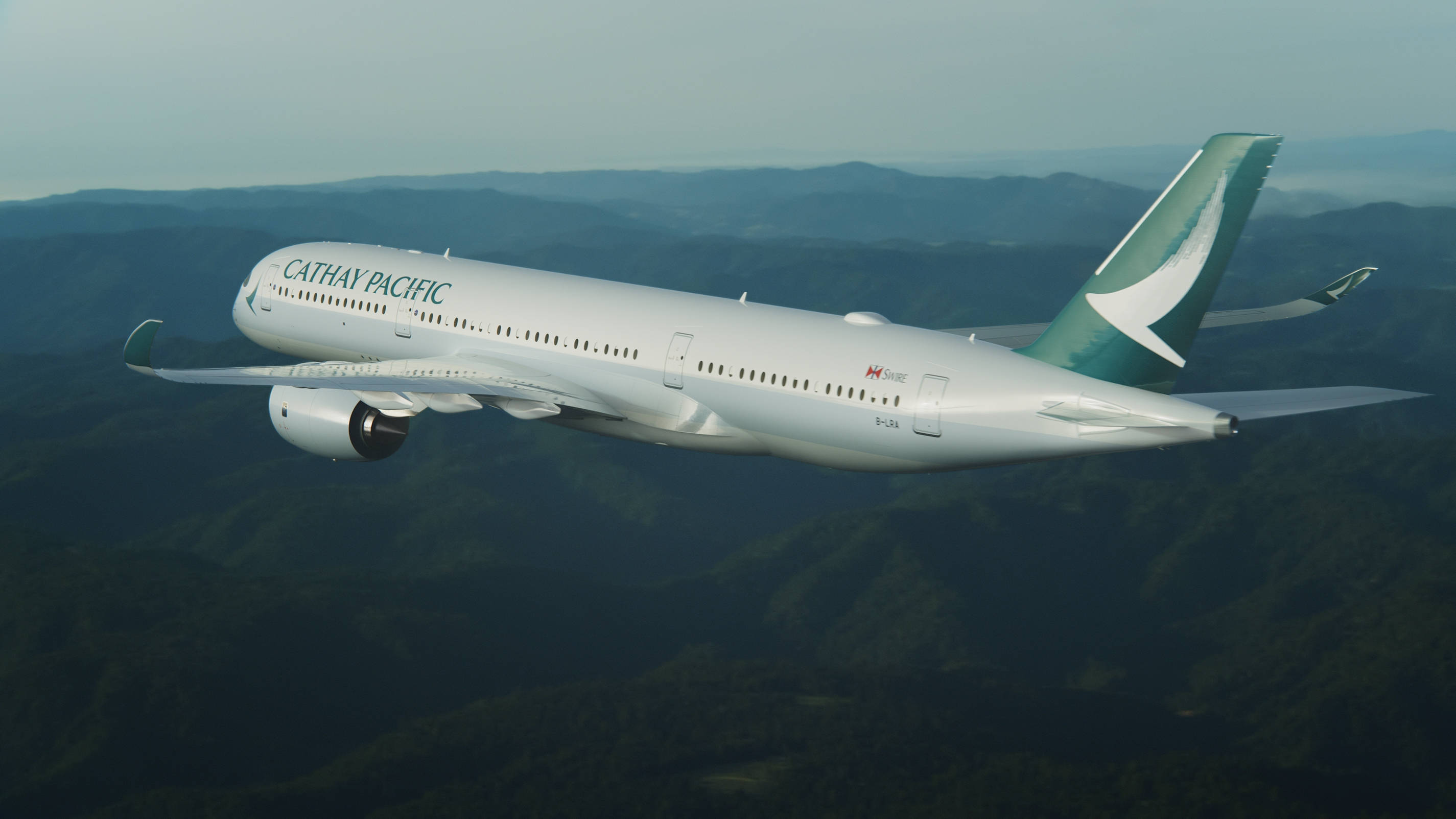 飛機 桌布 壁紙 — 國泰航空飛機在柔和天空下優雅地翱翔於翠綠山脈之上。