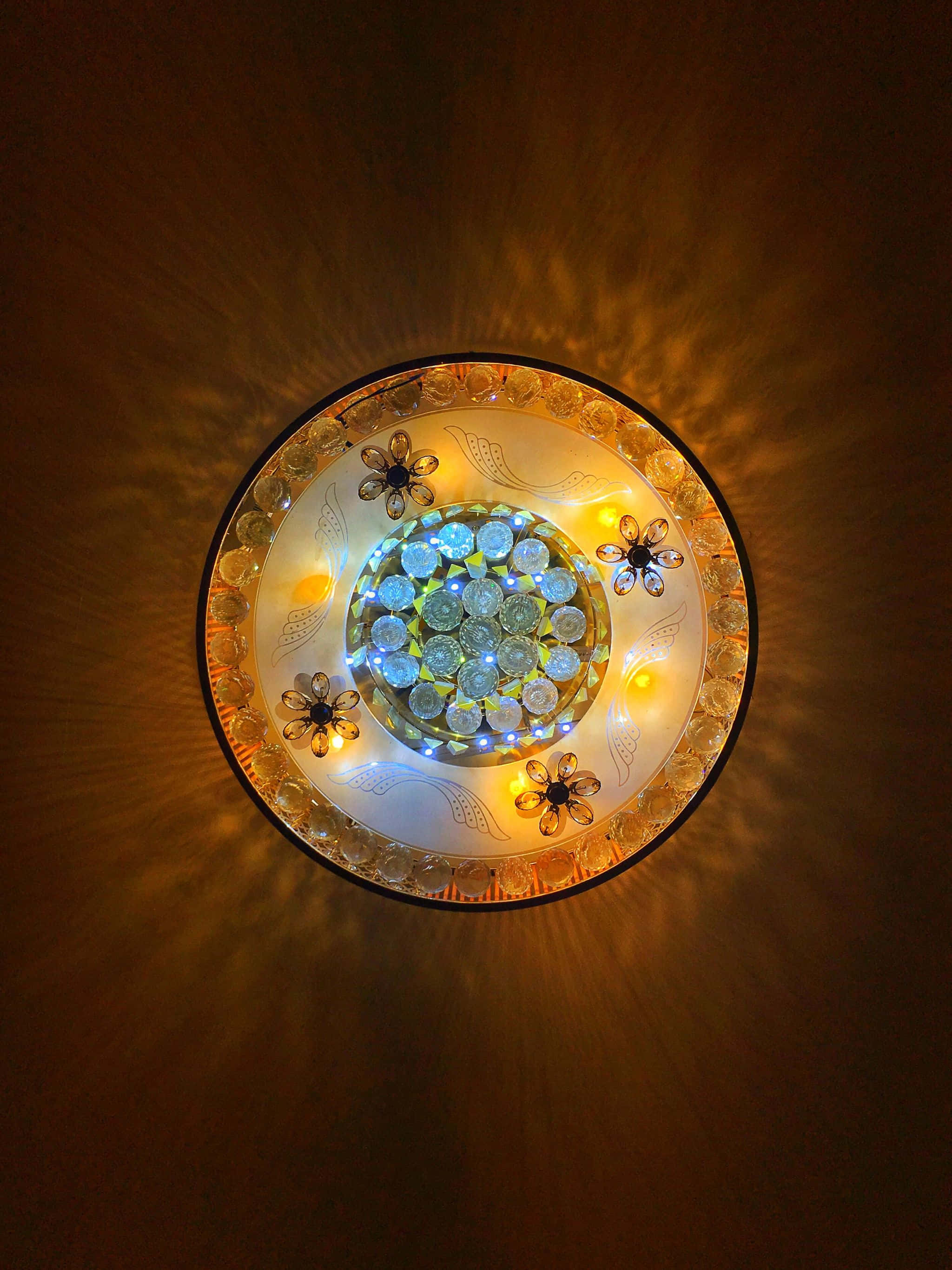 2048x2732 桌布 壁紙 — 圓形天花板燈具，帶有花卉水晶裝飾和發光的藍色及黃色燈泡，在深色牆壁上投射出溫暖的光線。