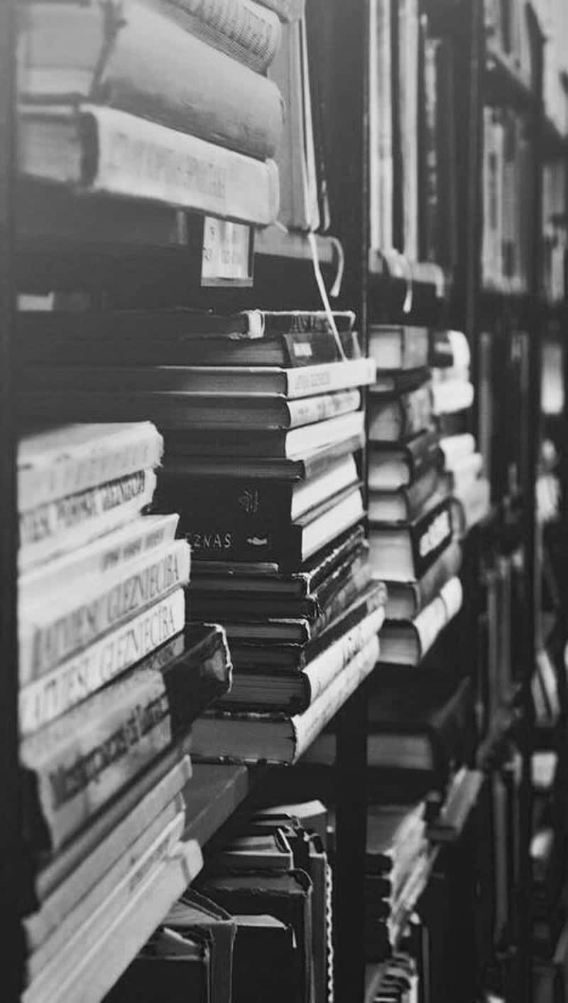 Black And White Book 桌布 壁紙 — 圖書館走廊中堆疊的書籍，以戲劇性的黑白風格捕捉，帶有深沉的陰影和對書脊的銳利對焦。
