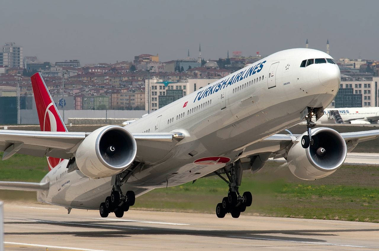 飛機 桌布 壁紙 — 土耳其航空波音777從跑道起飛，朦朧背景中可見城市景觀和宣禮塔。