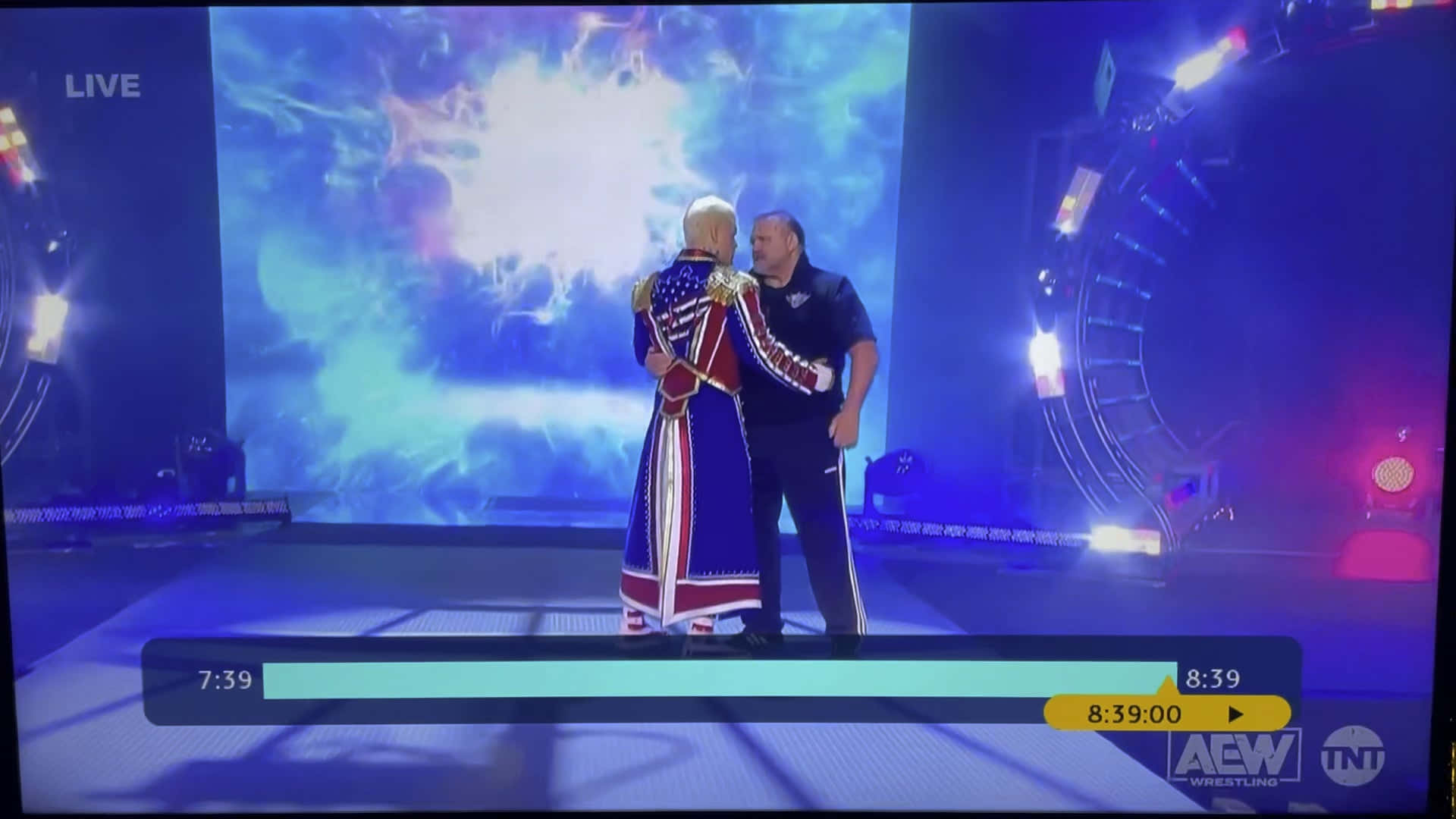Cody Rhodes 桌布 壁紙 — 在AEW摔角直播中，兩名摔角手在燈光明亮的舞台上擁抱，背景是宇宙星空。