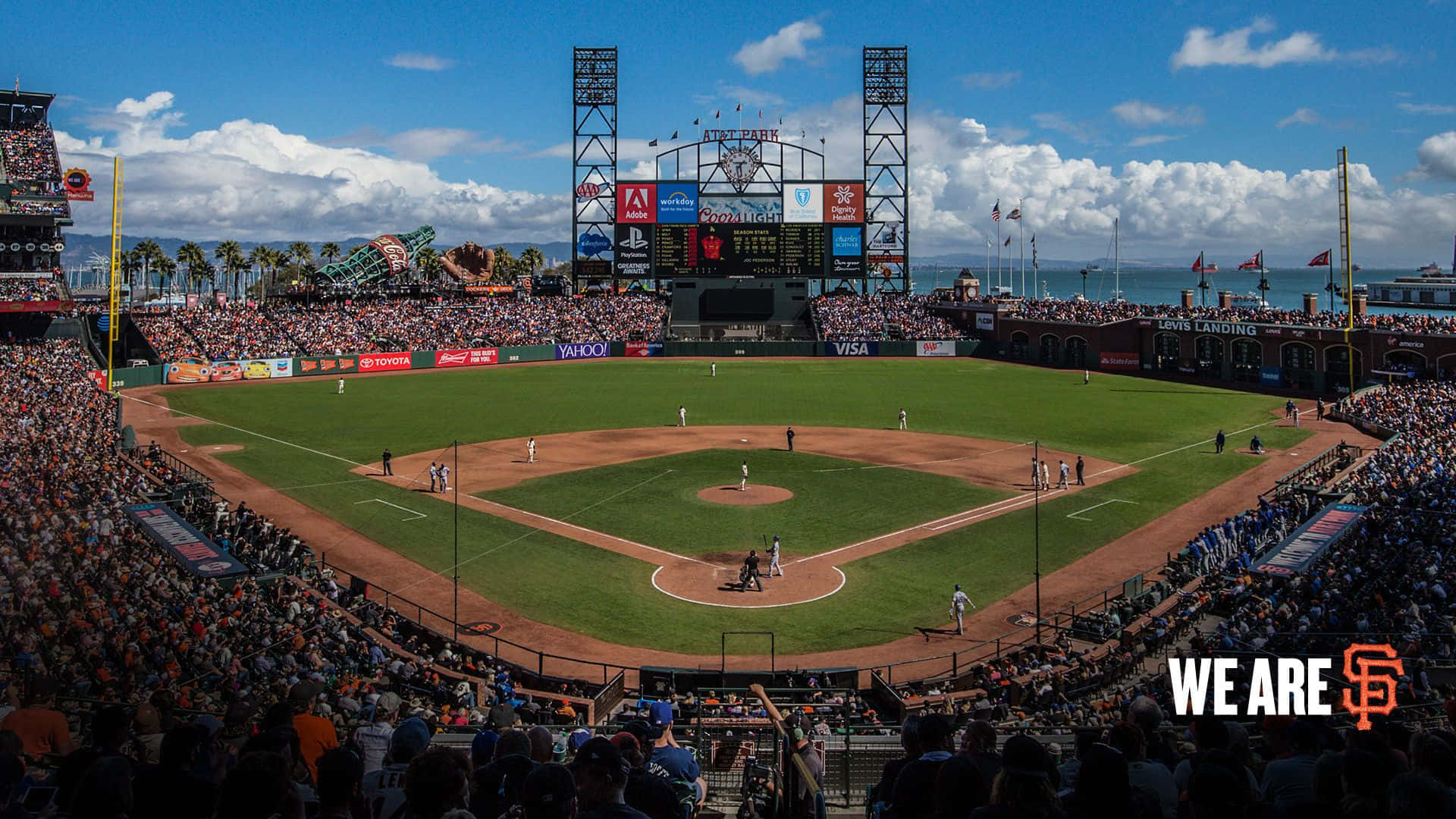 Baseball Stadium 桌布 壁紙 — 在AT&T公園，一場充滿活力的棒球比賽在明亮的藍天白雲下展開，周圍是歡呼的球迷和海洋景觀。