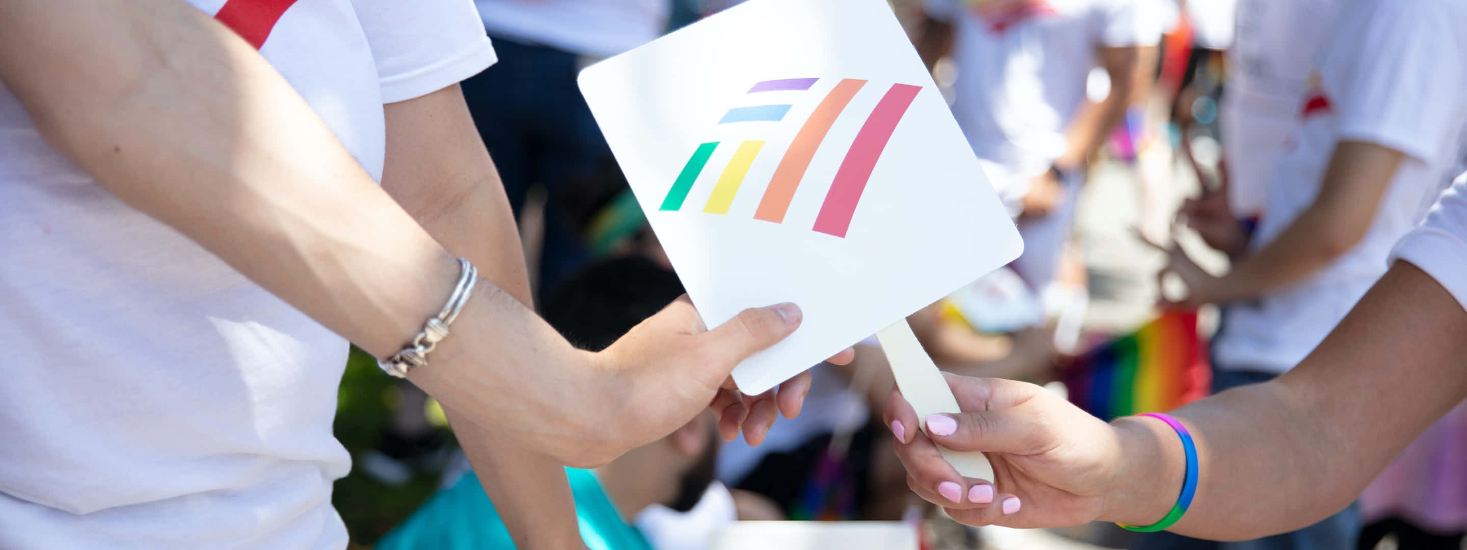 美國銀行 桌布 壁紙 — 在一個充滿活力的戶外Pride活動中，兩隻手交換一把帶有彩虹條紋的白色扇子。