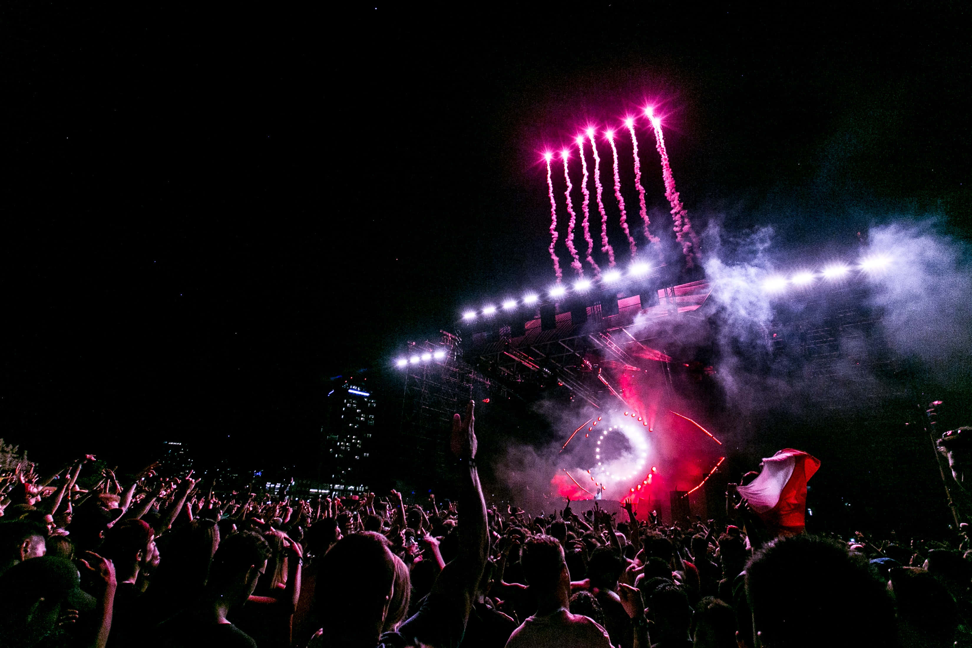 music festival 桌布 壁紙 — 在一個大型戶外音樂會上，夜晚的喧鬧人群在耀眼的粉紅色煙火和明亮的舞台燈光下舉起雙手。