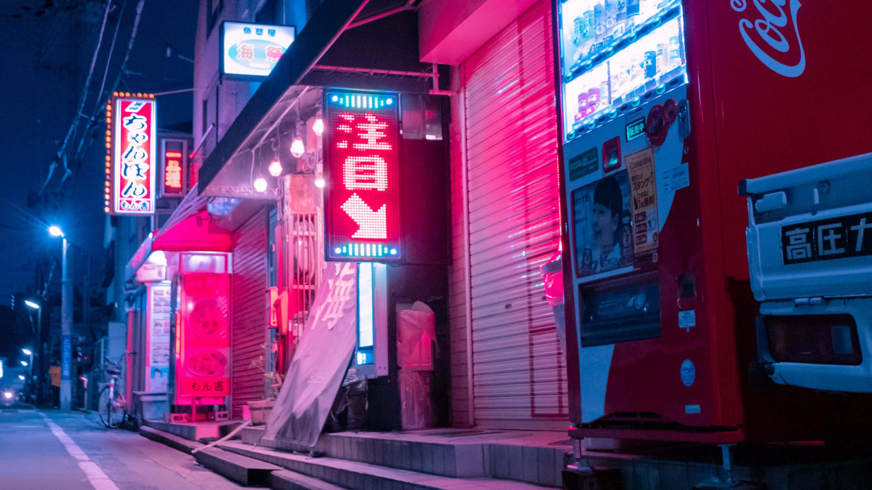 日式粉紅 桌布 壁紙 — 在一個寧靜的日本夜晚街道上，鮮豔的霓虹燈牌閃爍著粉紅色和藍色光芒，旁邊有一部紅色自動販賣機和關閉的商店。