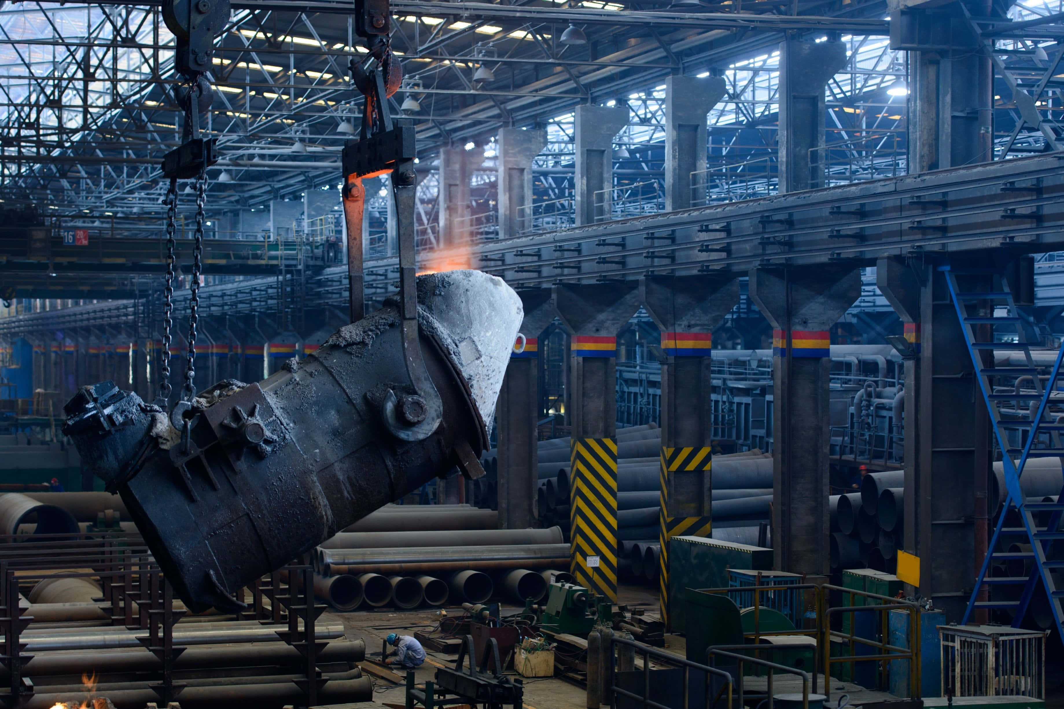 Manufacturing 桌布 壁紙 — 在一個廣闊的工業鋼鐵廠中，有暴露的鋼樑和管道，一個發光的熔融金屬澆注桶被起重機吊起。
