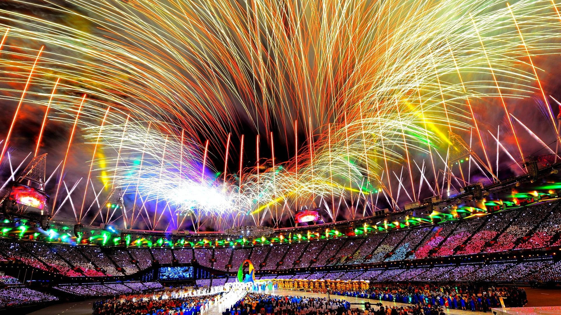 Olympics 桌布 壁紙 — 在一個擠滿觀眾的體育場上空，一場壯觀的煙火表演在夜間活動中綻放，伴隨著鮮豔的燈光和歡呼的人群。