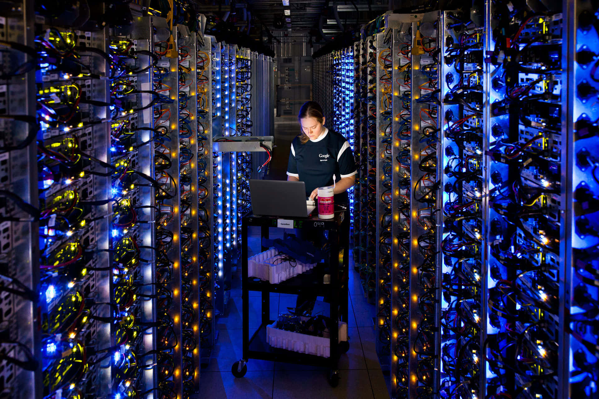 data 桌布 壁紙 — 在一個高科技資料中心，技術人員在散發藍色和黃色光芒的高聳伺服器機架之間使用筆記型電腦工作。