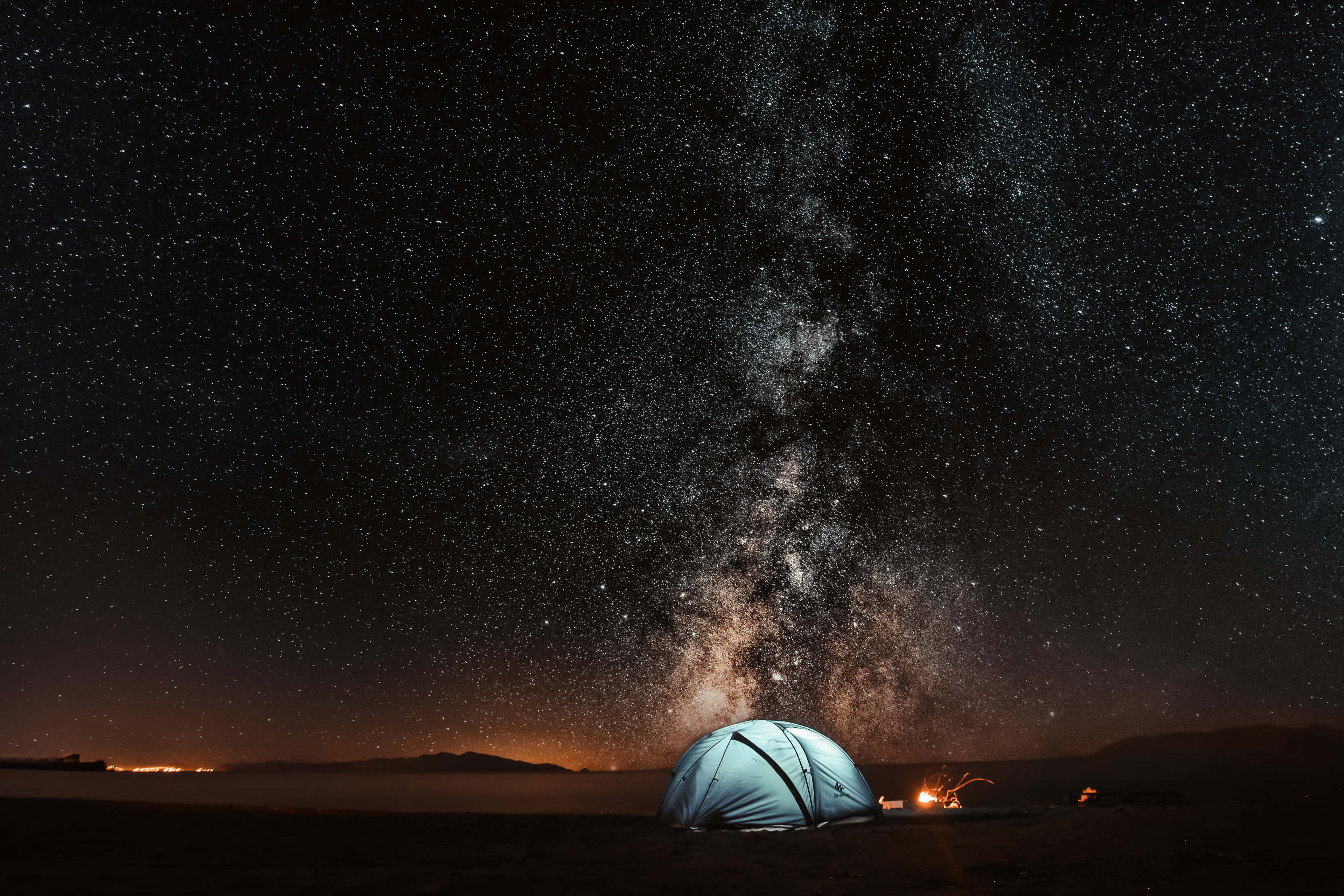 Camping 桌布 壁紙 — 在令人嘆為觀止的星空下，一個發光的藍色帳篷，銀河在頭頂拱起，地平線上有遠處的城市燈光。