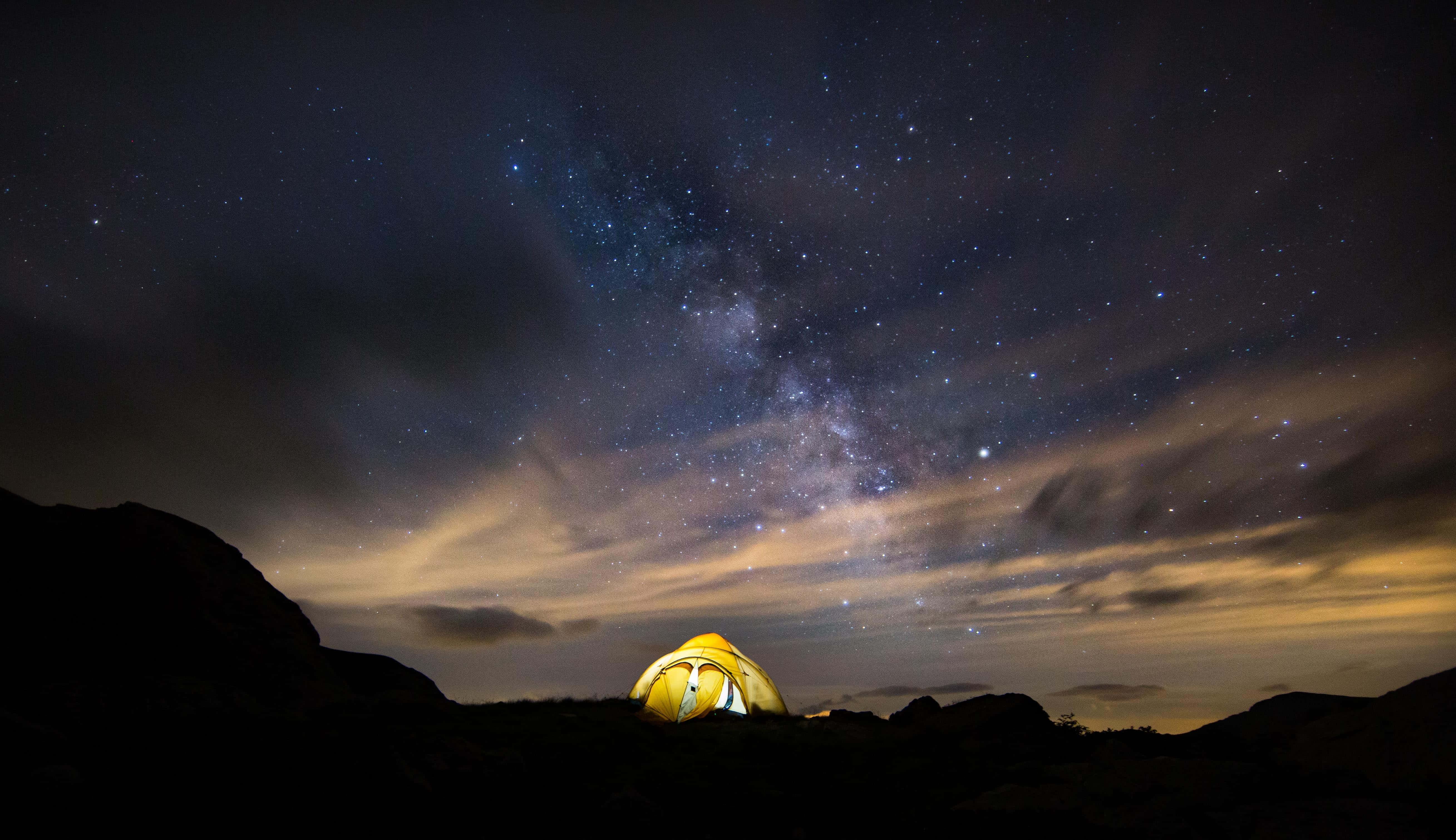 Camping 桌布 壁紙 — 在令人驚嘆的星空下，一個發光的黃色帳篷坐落在黑暗的岩石地形中，銀河在頭頂拱起。