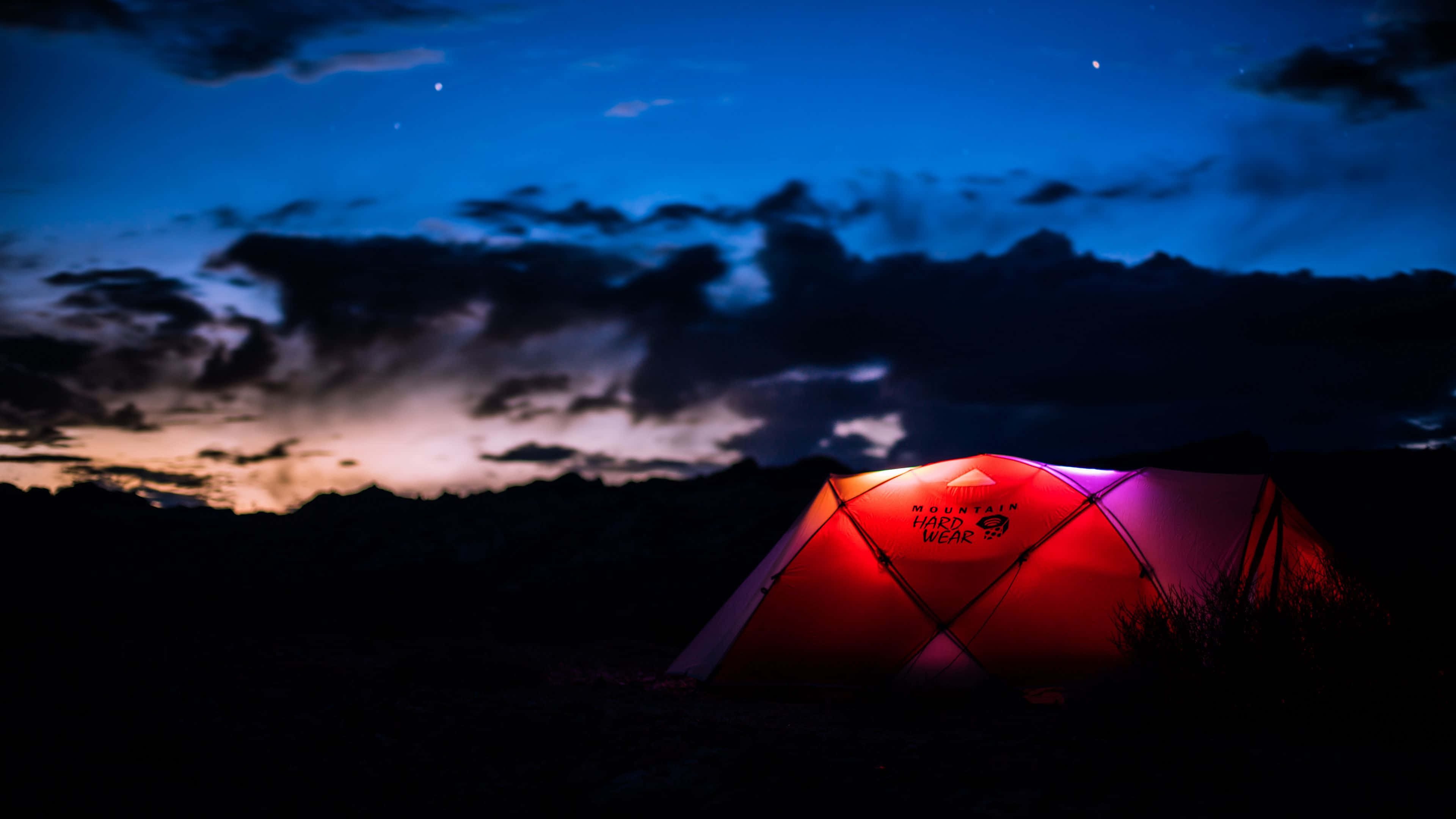 Camping 桌布 壁紙 — 在佈滿紫色和橙色雲朵的星空暮色下，一頂發光的紅色帳篷照亮了黑暗的山間營地。