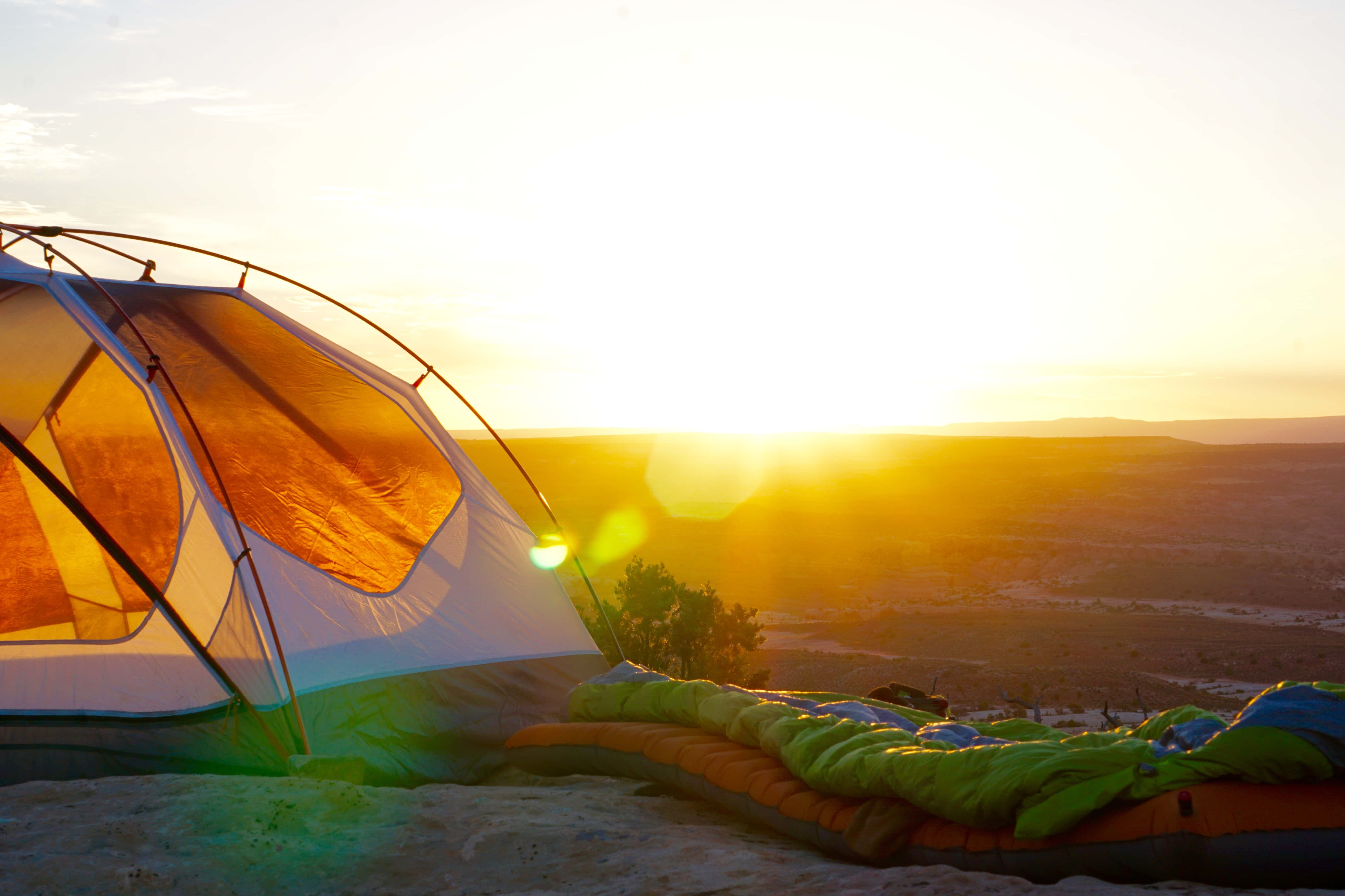 Camping 桌布 壁紙 — 在俯瞰廣闊峽谷的沙漠懸崖上，帳篷和睡袋在日出時溫暖地發光。