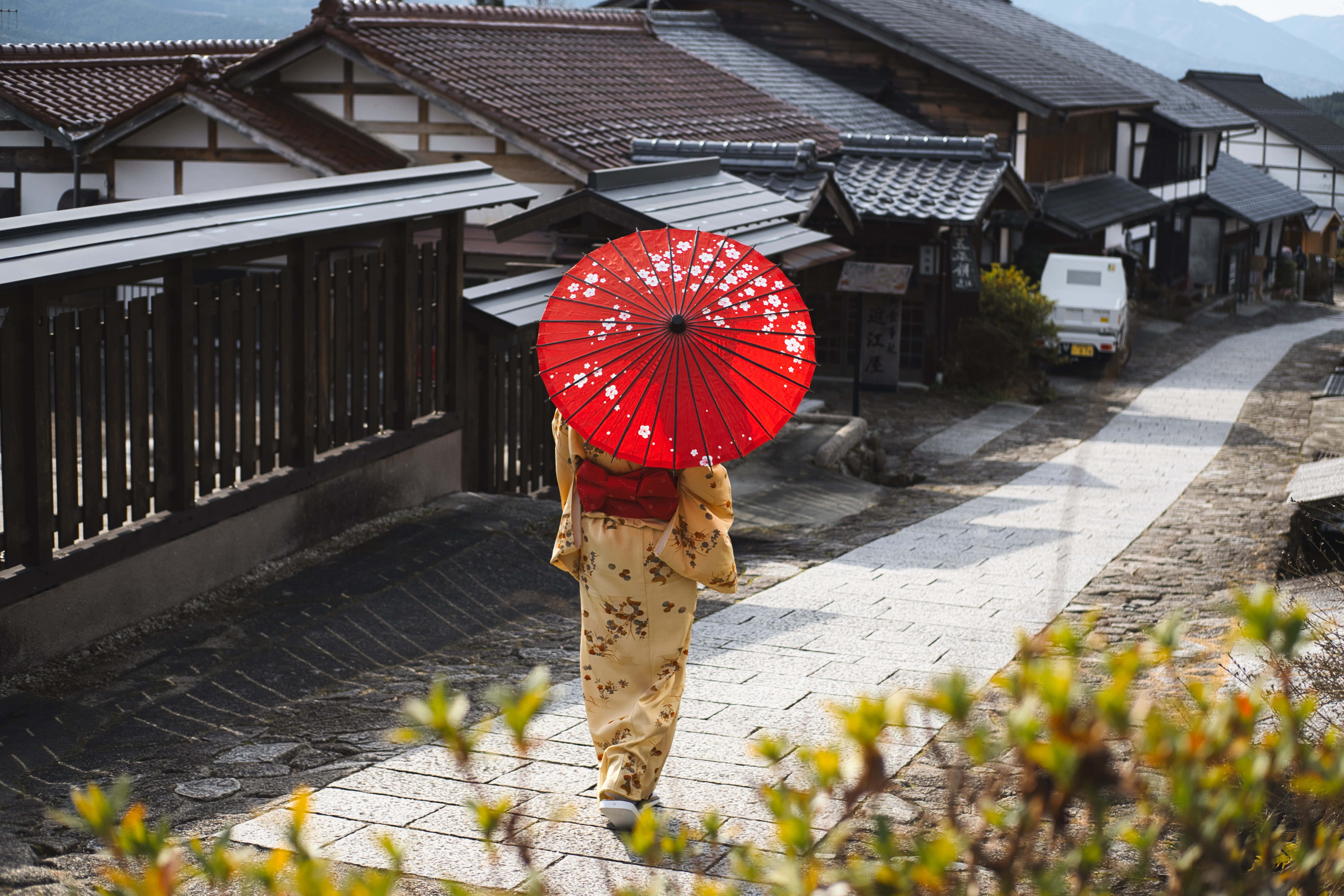 Asian 桌布 壁紙 — 在傳統日本村莊中，一位穿著花卉和服的人撐著紅色櫻花傘沿著石徑走遠。