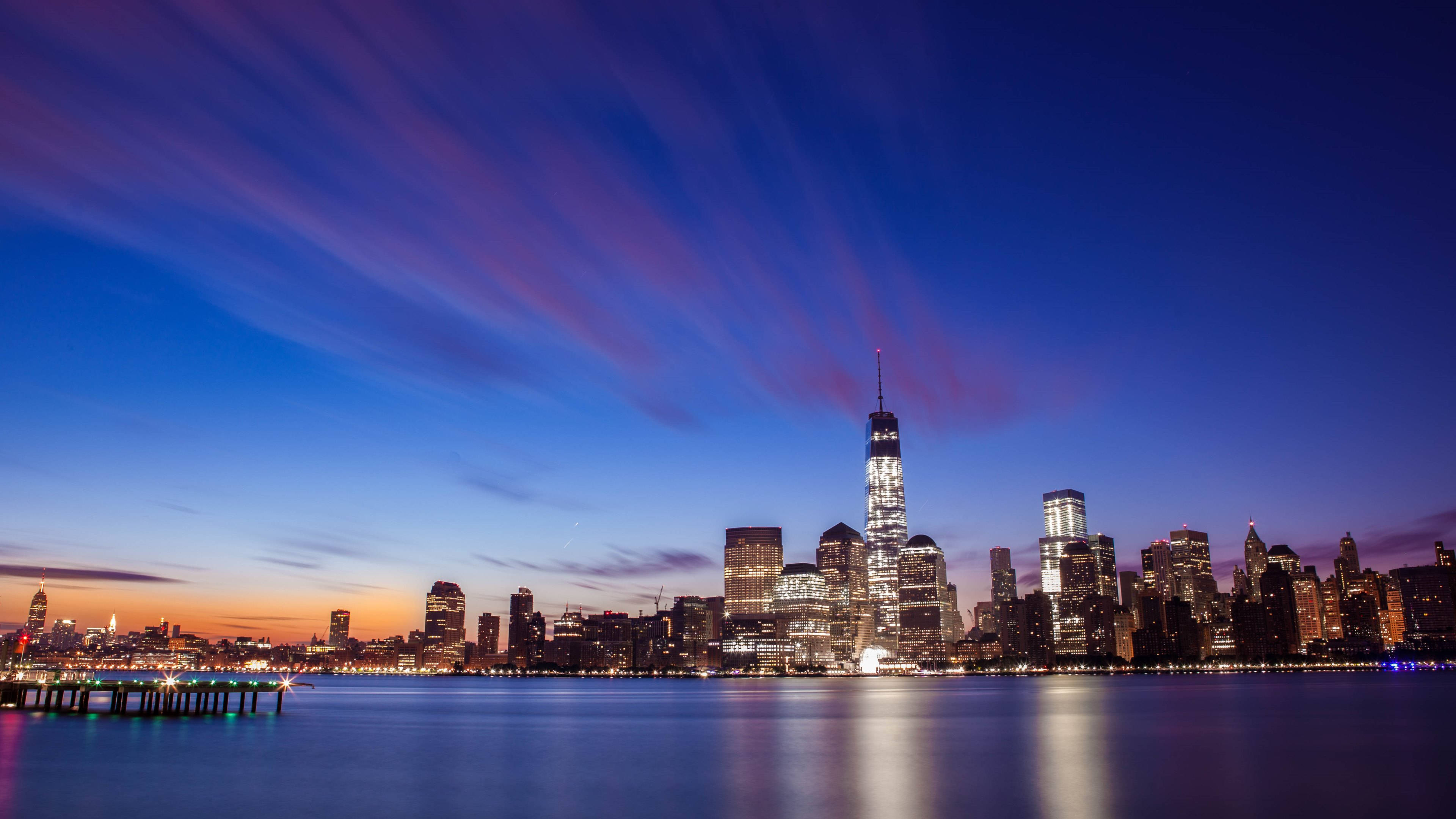 New York City Desktop 桌布 壁紙 — 在充滿活力的紫色和橙色天空下，紐約市天際線的壯麗黃昏景色，明亮的摩天大樓倒映在平靜的水面上。