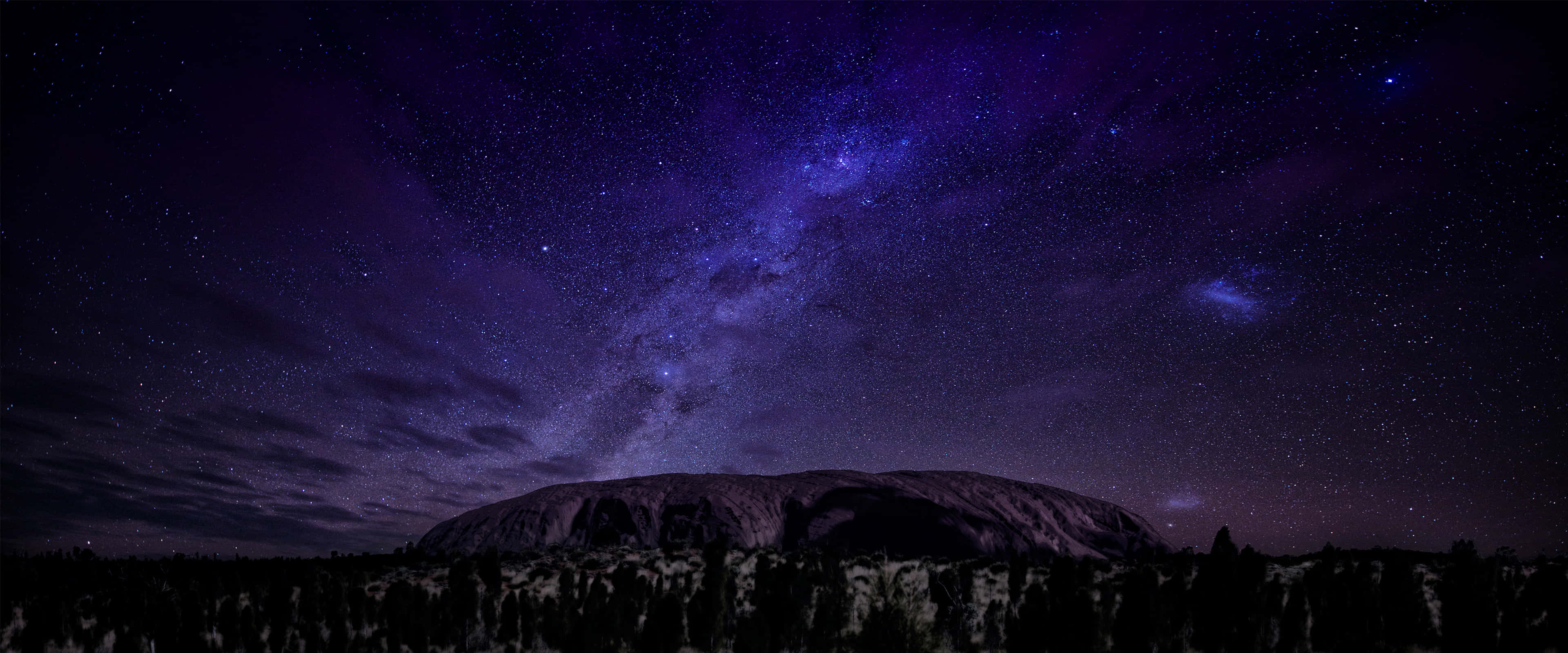 Apple 3840x1600 桌布 壁紙 — 在充滿活力的紫色星空下，一個雄偉的黑色岩石輪廓，銀河在上方閃耀。
