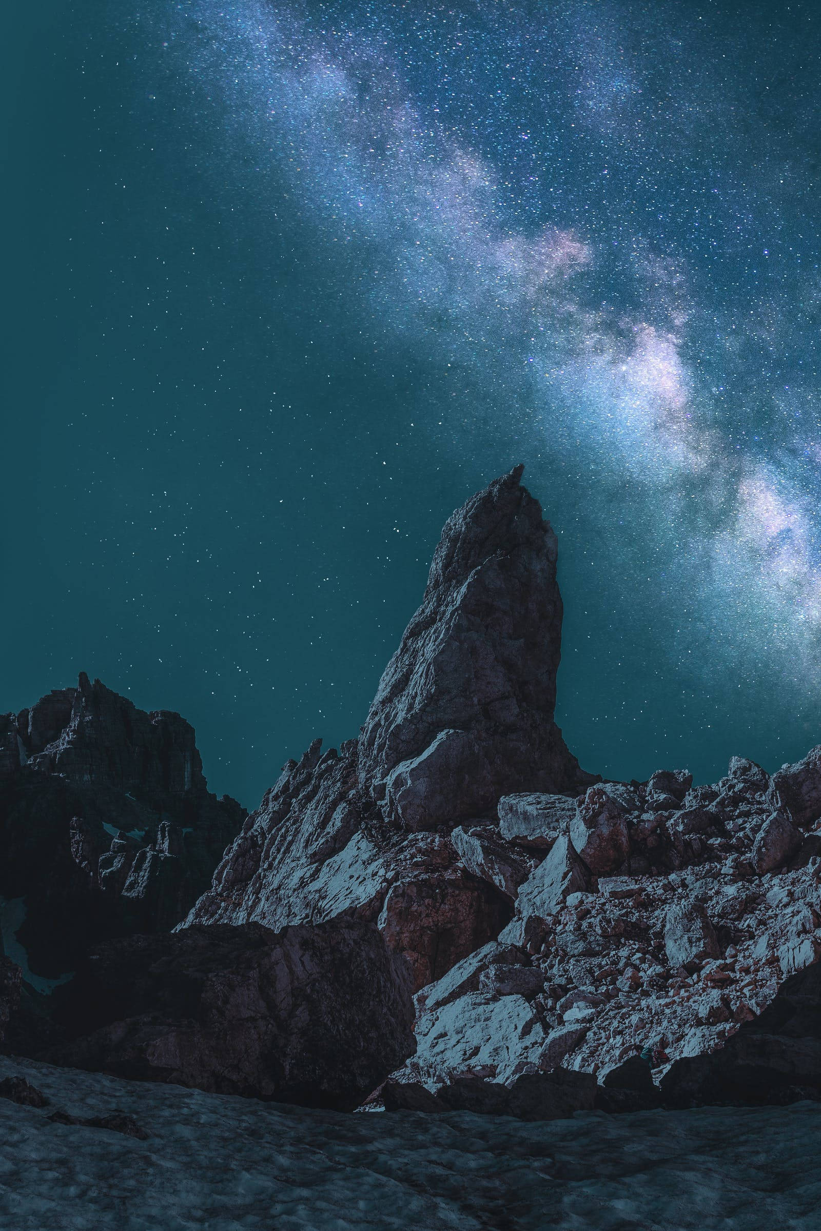 太空 iPhone 桌布 壁紙 — 在充滿銀河系閃爍星星的鮮豔藍綠色天空下，一座崎嶇的山峰聳立。