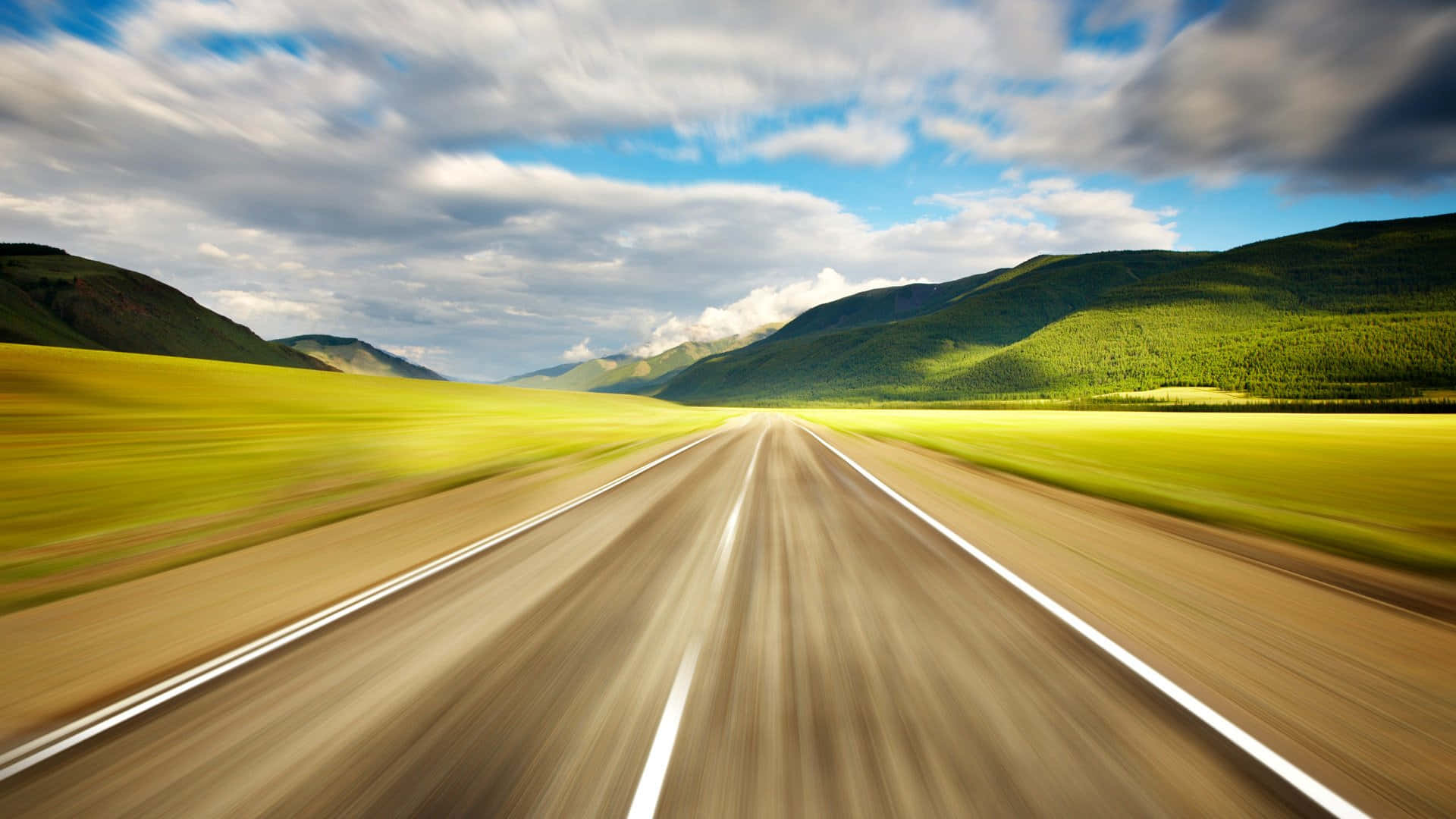 highway 桌布 壁紙 — 在動態多雲的天空下，一條筆直的道路兩旁是充滿活力的黃色田野和綠色山脈，高速行駛的模糊視野。