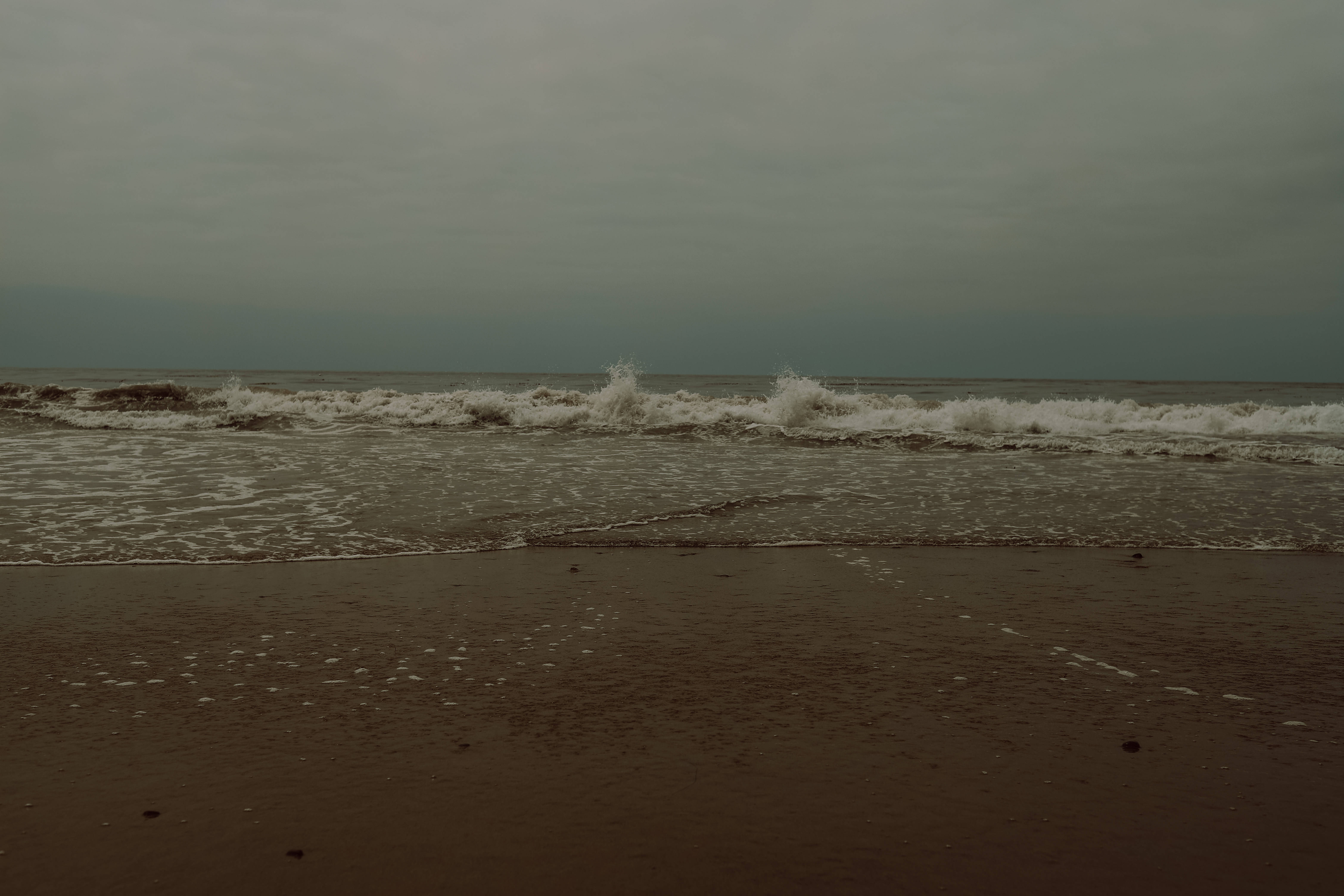Aesthetic For Laptop 桌布 壁紙 — 在厚重的陰天下，海浪拍打著潮濕的沙灘，呈現柔和的棕色和灰色色調。