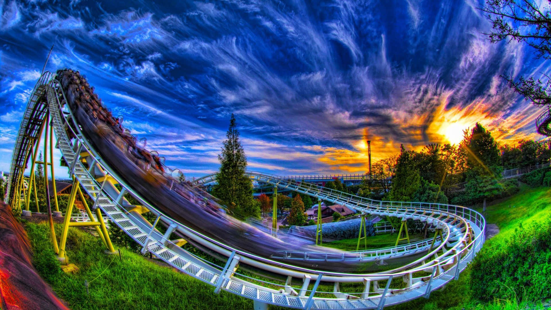 Roller Coaster 桌布 壁紙 — 在壯麗的日落天空下，雲朵翻騰，一輛雲霄飛車沿著陡峭的軌道疾馳而下。