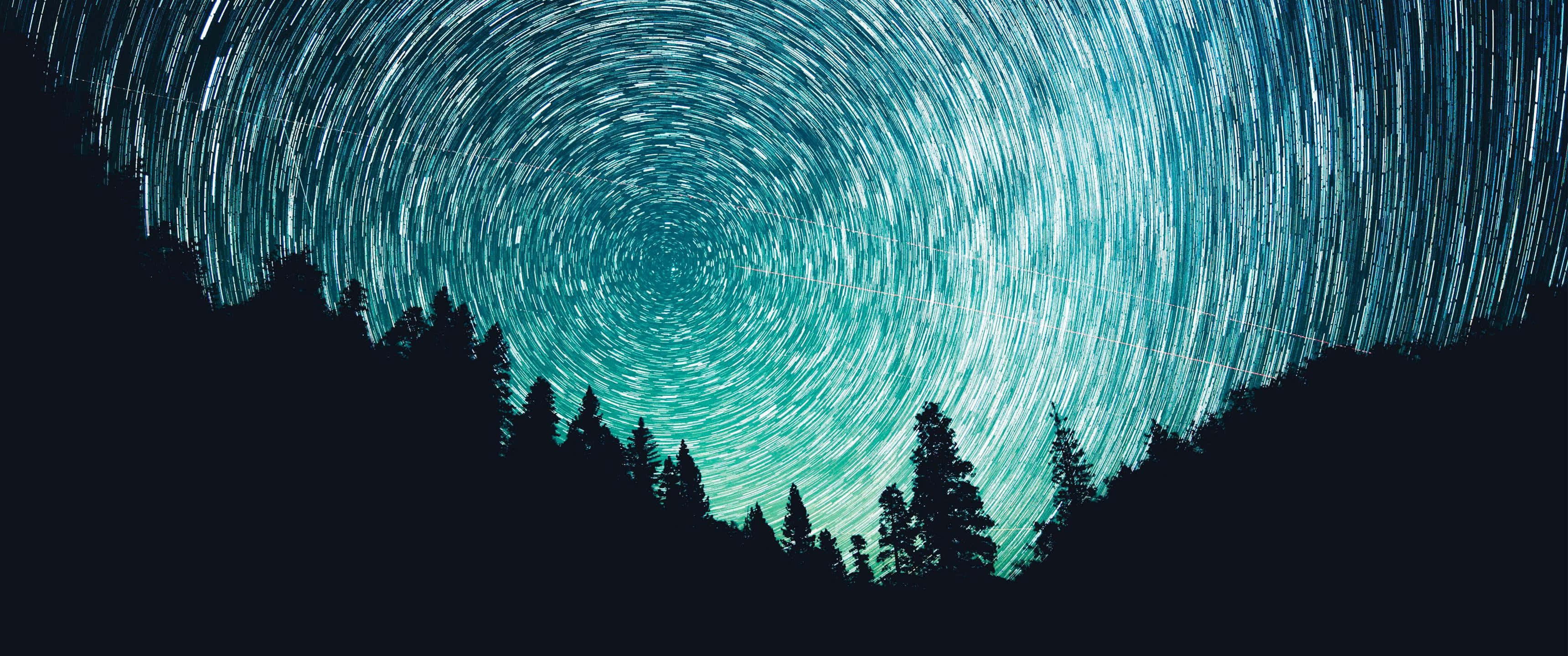 Ultra Wide 3440x1440 Space 桌布 壁紙 — 在夜空下，深色松樹林剪影上方，青綠色光芒中盤旋的星軌。