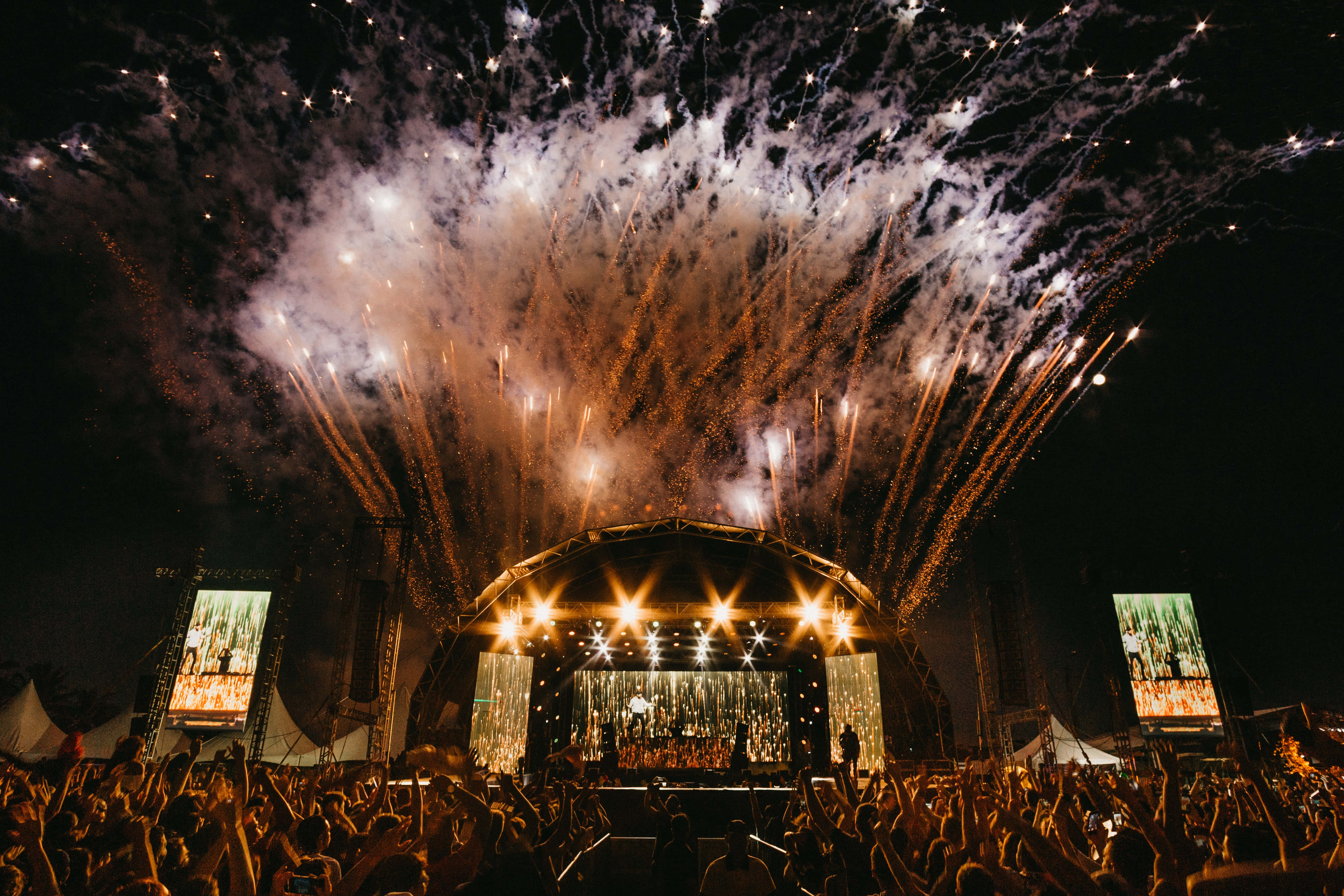 music festival 桌布 壁紙 — 在夜間明亮照明的音樂會舞台上，金色煙火璀璨爆發，一群喧鬧的人群在下方歡呼。