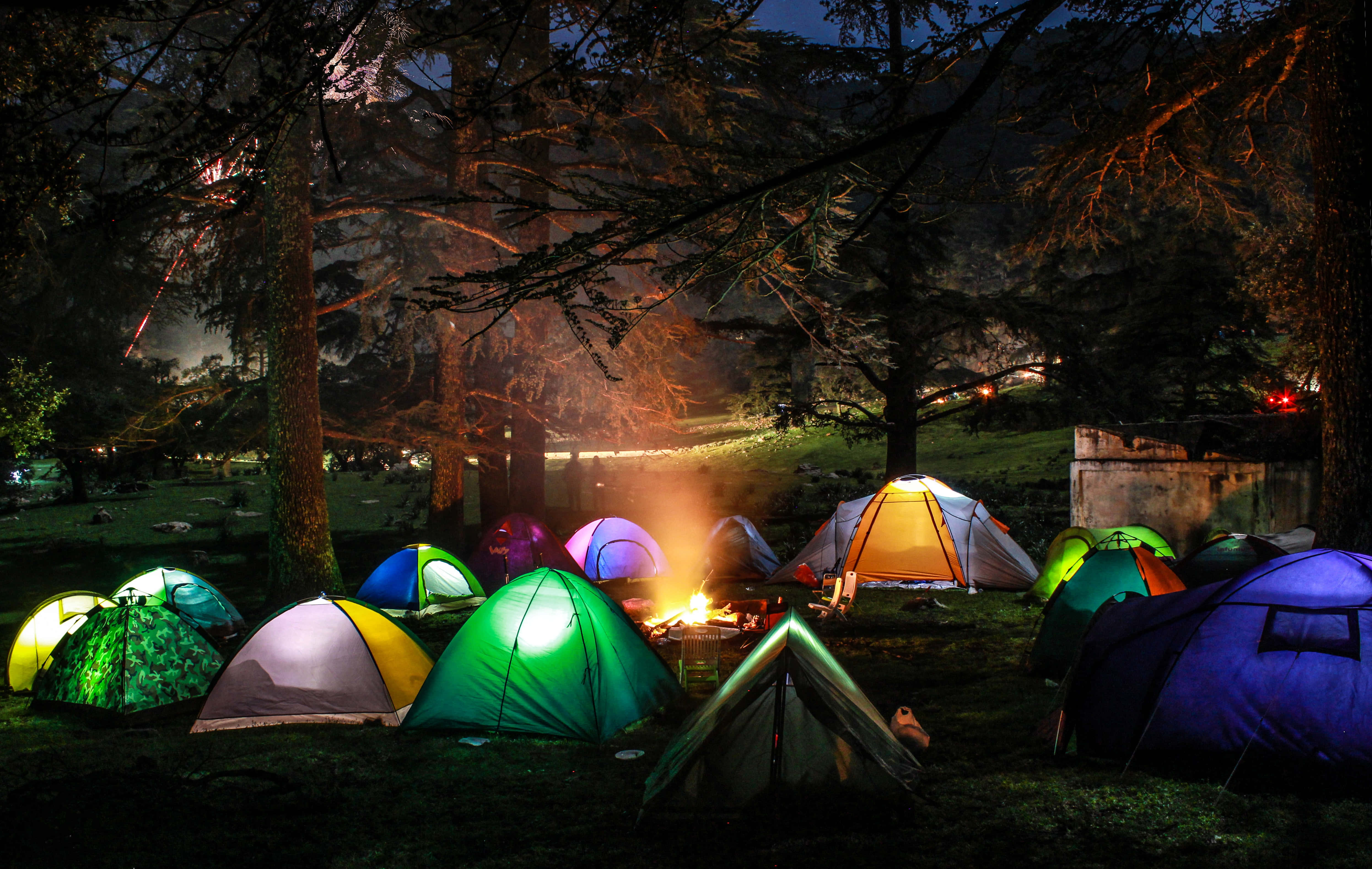 Camping 桌布 壁紙 — 在夜間森林營地中，色彩繽紛的發光帳篷圍繞著溫暖的營火，位於黑暗的樹木下。