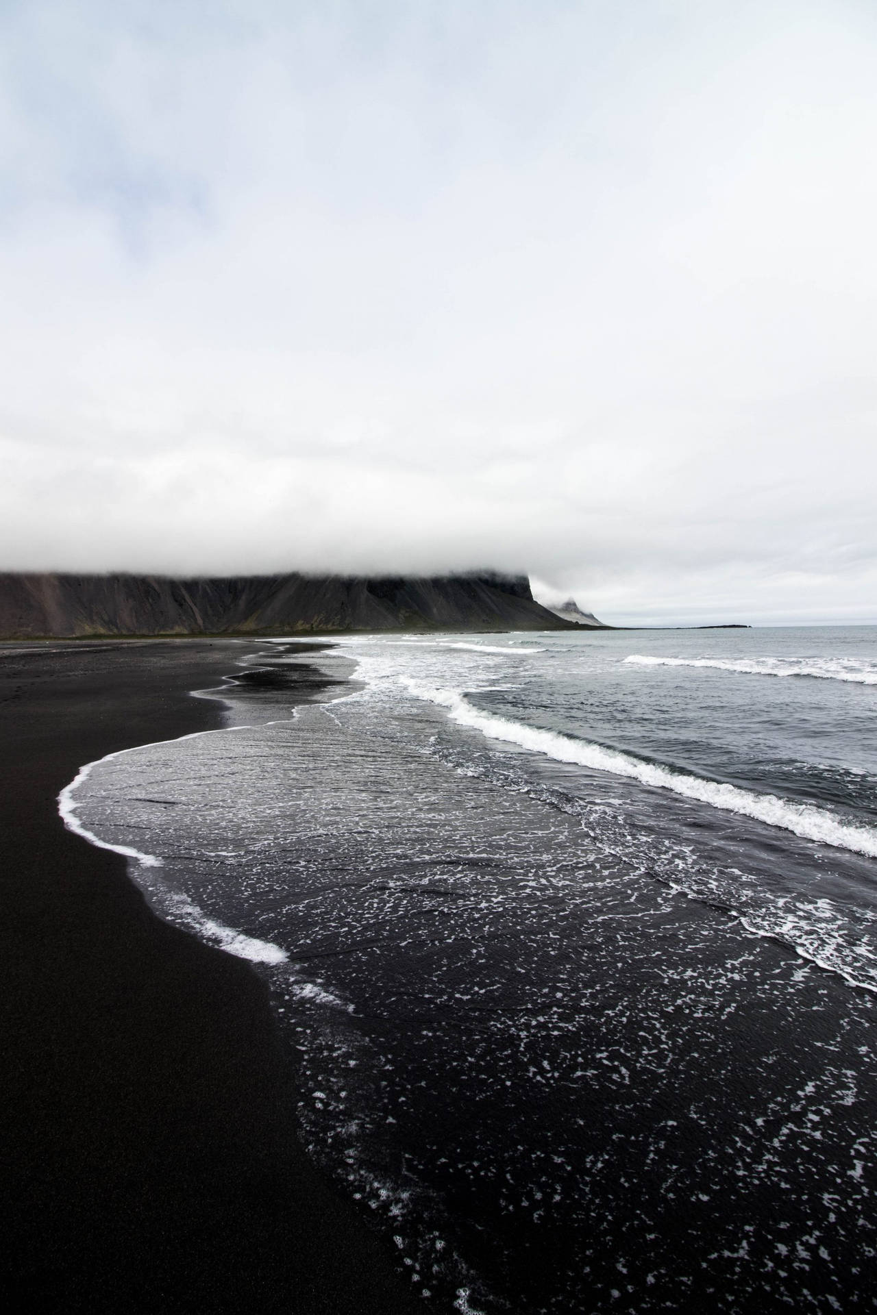 iPhone 美學 桌布 壁紙 — 在密雲天空下，海浪拍打著黑色的火山沙灘，霧氣籠罩著遠處的懸崖。