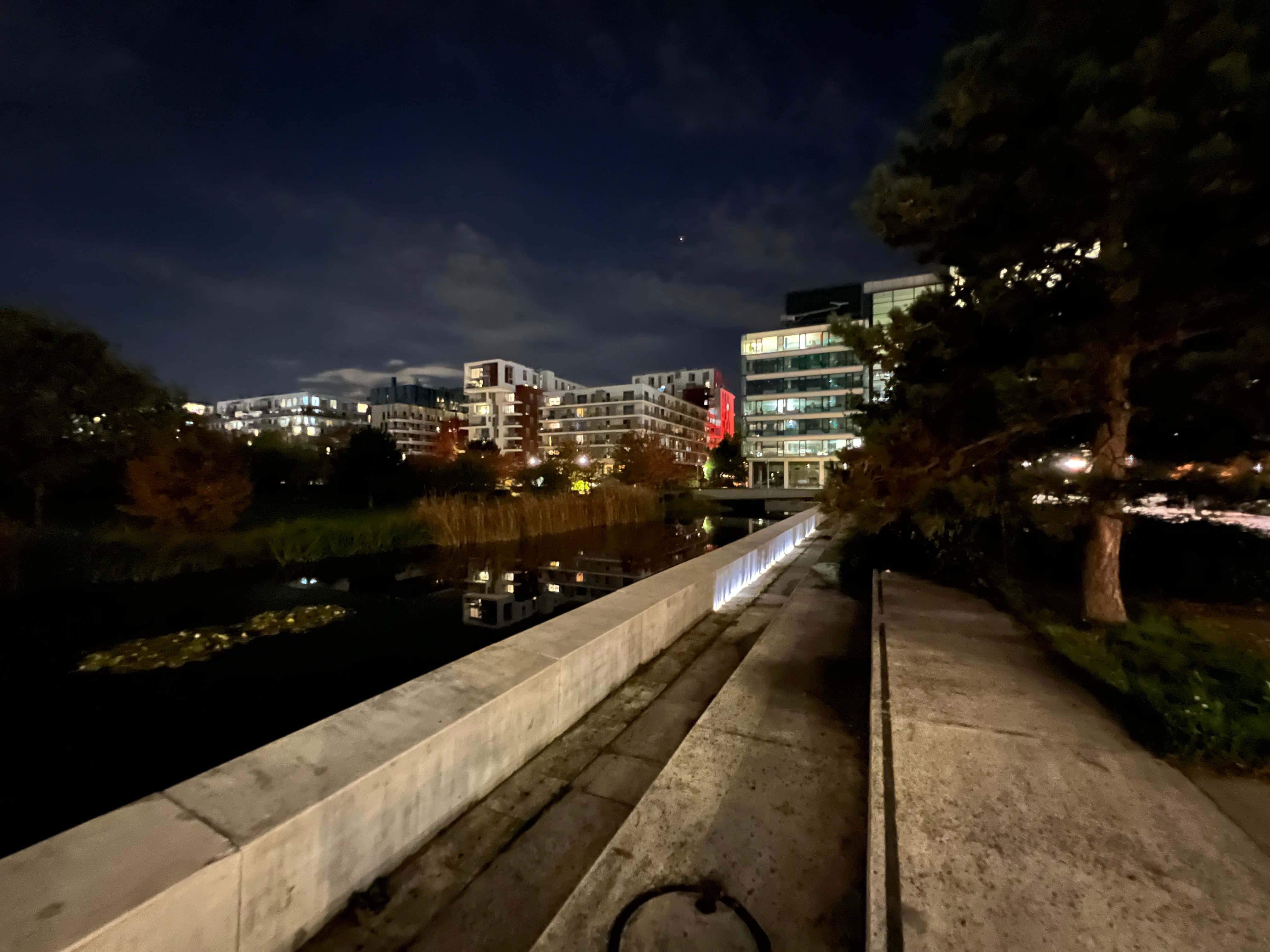 Night Mode 桌布 壁紙 — 在寧靜的夜晚沿著混凝土小徑散步，旁邊是平靜的運河，倒映著發光的城市建築和樹木，天空一片漆黑。