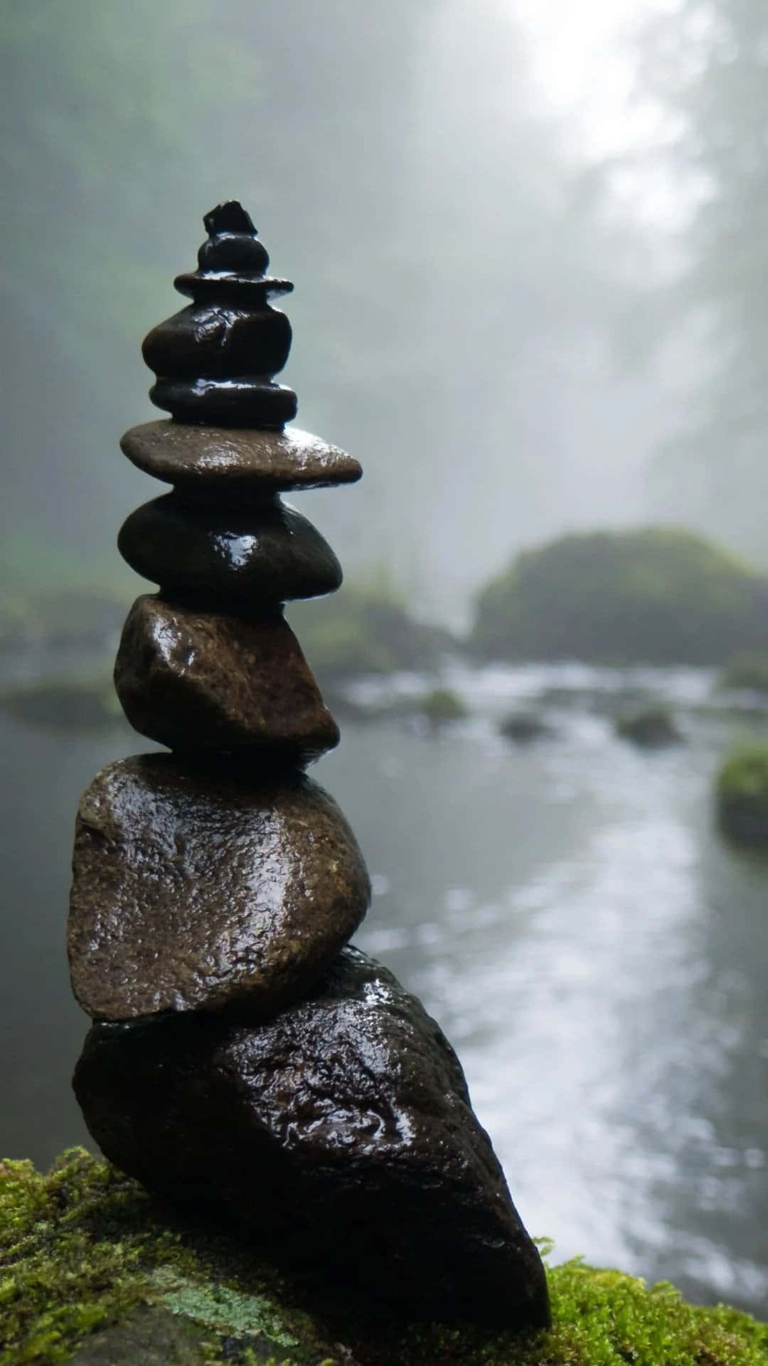 禪 桌布 壁紙 — 在寧靜的森林中，一堆濕漉漉的河石精巧地平衡在長滿苔蘚的岩石上，旁邊是霧氣瀰漫、流淌的小溪。
