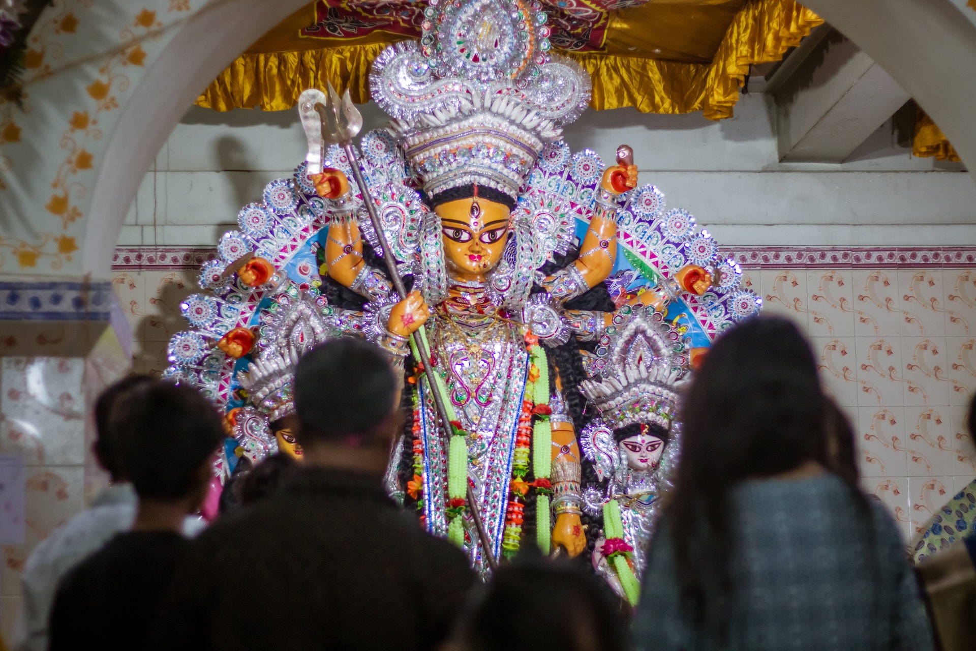 Hindu Temple 桌布 壁紙 — 在寺廟環境中，從信徒背後看到的色彩鮮豔、裝飾精美的杜爾迦神像，擁有多臂和武器。