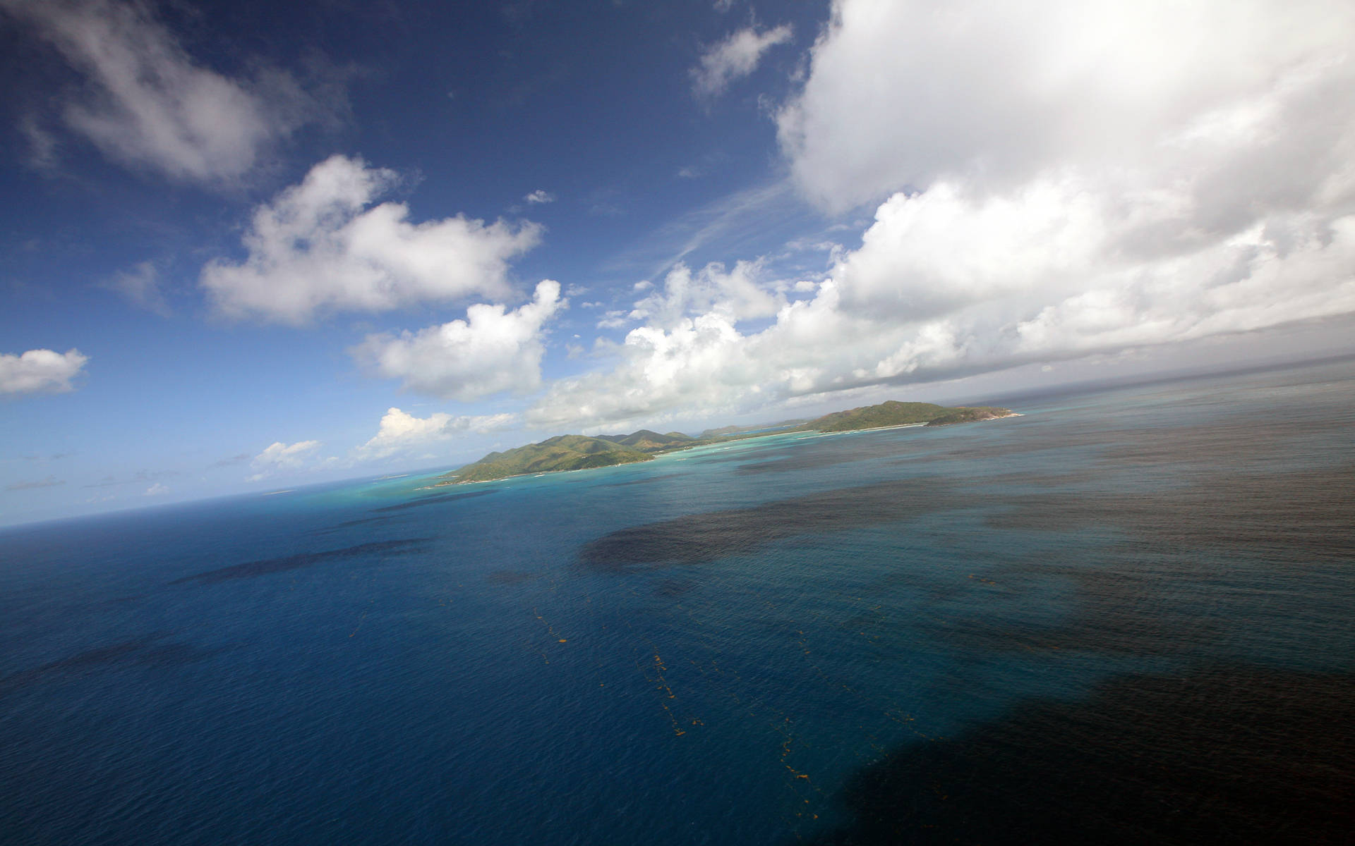 Seychelles 桌布 壁紙 — 在局部多雲的天空下，一個鬱鬱蔥蔥的綠色島嶼被充滿活力的藍綠色和深藍色海洋包圍的航拍視圖。