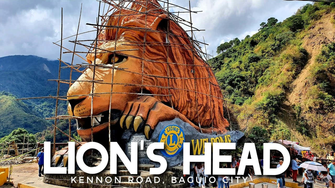 Lion Head 桌布 壁紙 — 在山路建造中的巨大獅子頭雕塑，背景有遊客和綠色山丘。