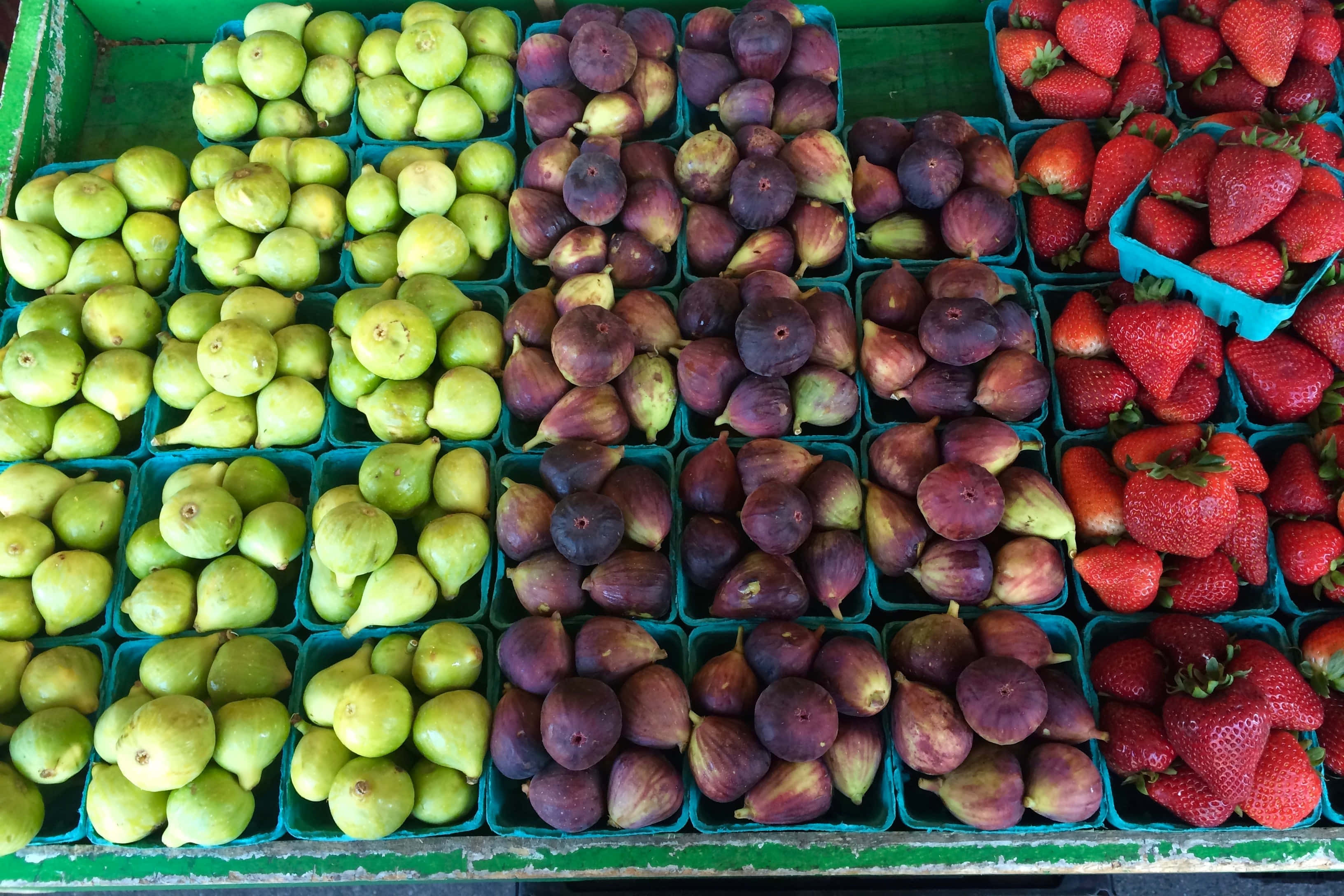 Farmers Market 桌布 壁紙 — 在市場攤位上，藍色塑膠容器中整齊排列著新鮮的綠色無花果、紫色無花果和紅色草莓，色彩繽紛。