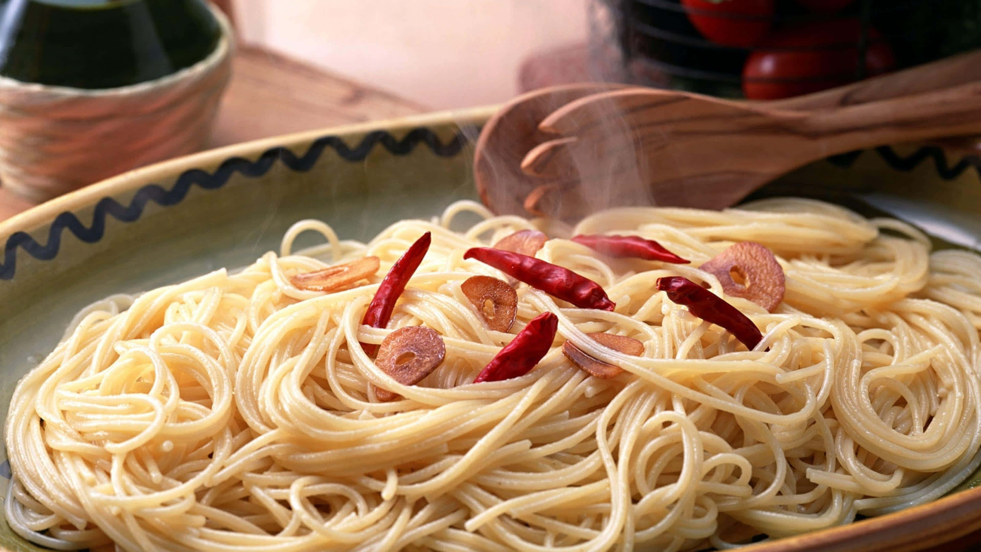 4K Pasta 桌布 壁紙 — 在帶有圖案的陶瓷碗中，冒著熱氣的義大利麵條上放著紅辣椒和蒜瓣。