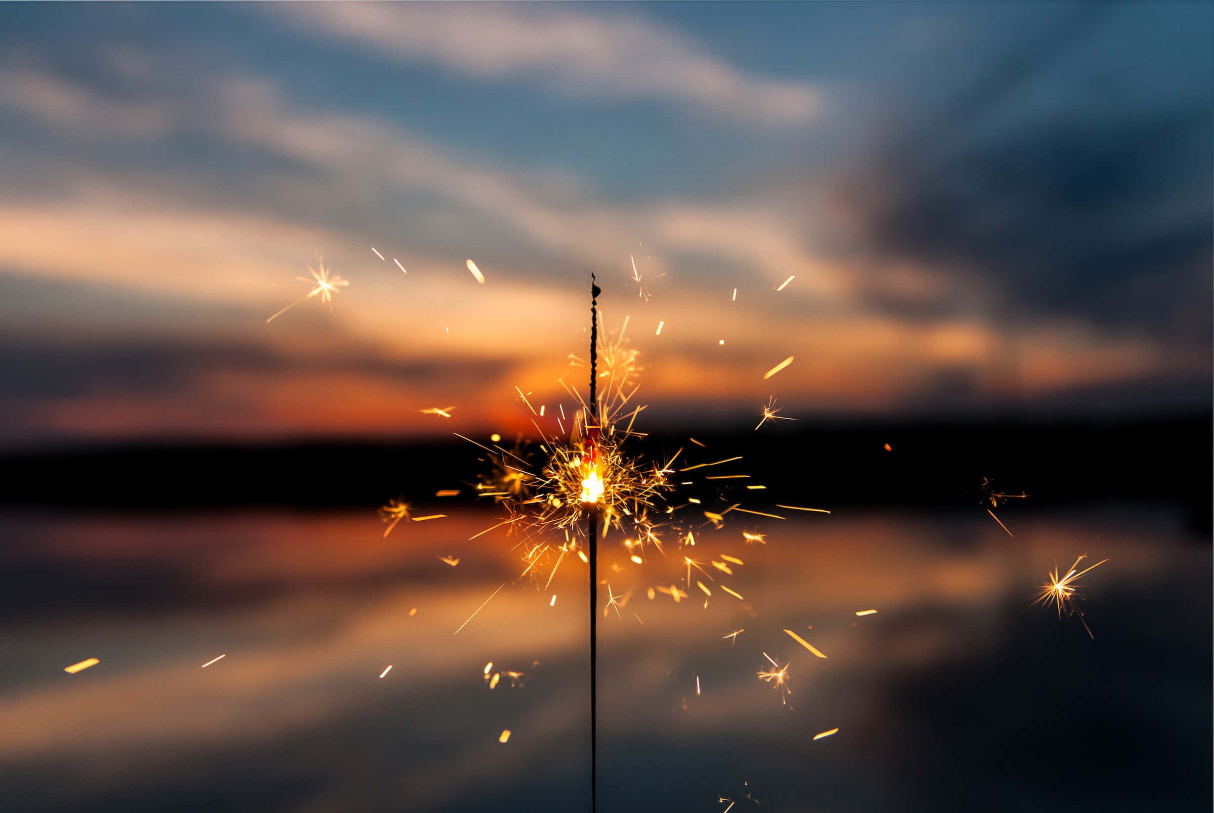 firework 桌布 壁紙 — 在平靜的水面上，閃爍的煙火在暮色天空中爆發出金色火花。