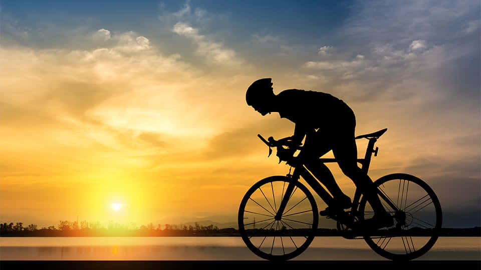 bike riding 桌布 壁紙 — 在平靜的水面上，騎自行車者的剪影正對著充滿活力的日落天空奮力踩踏。